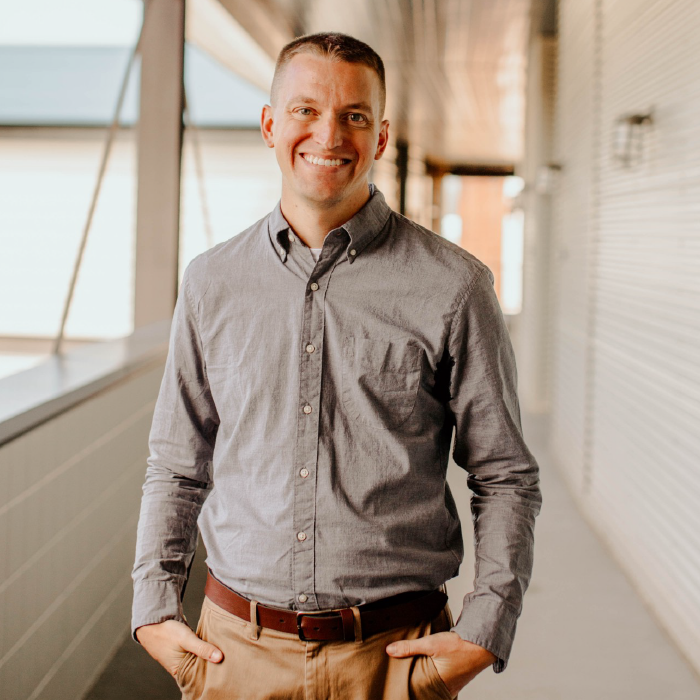 Smiling man in gray button-up shirt and khakis standing in a bright hallway.