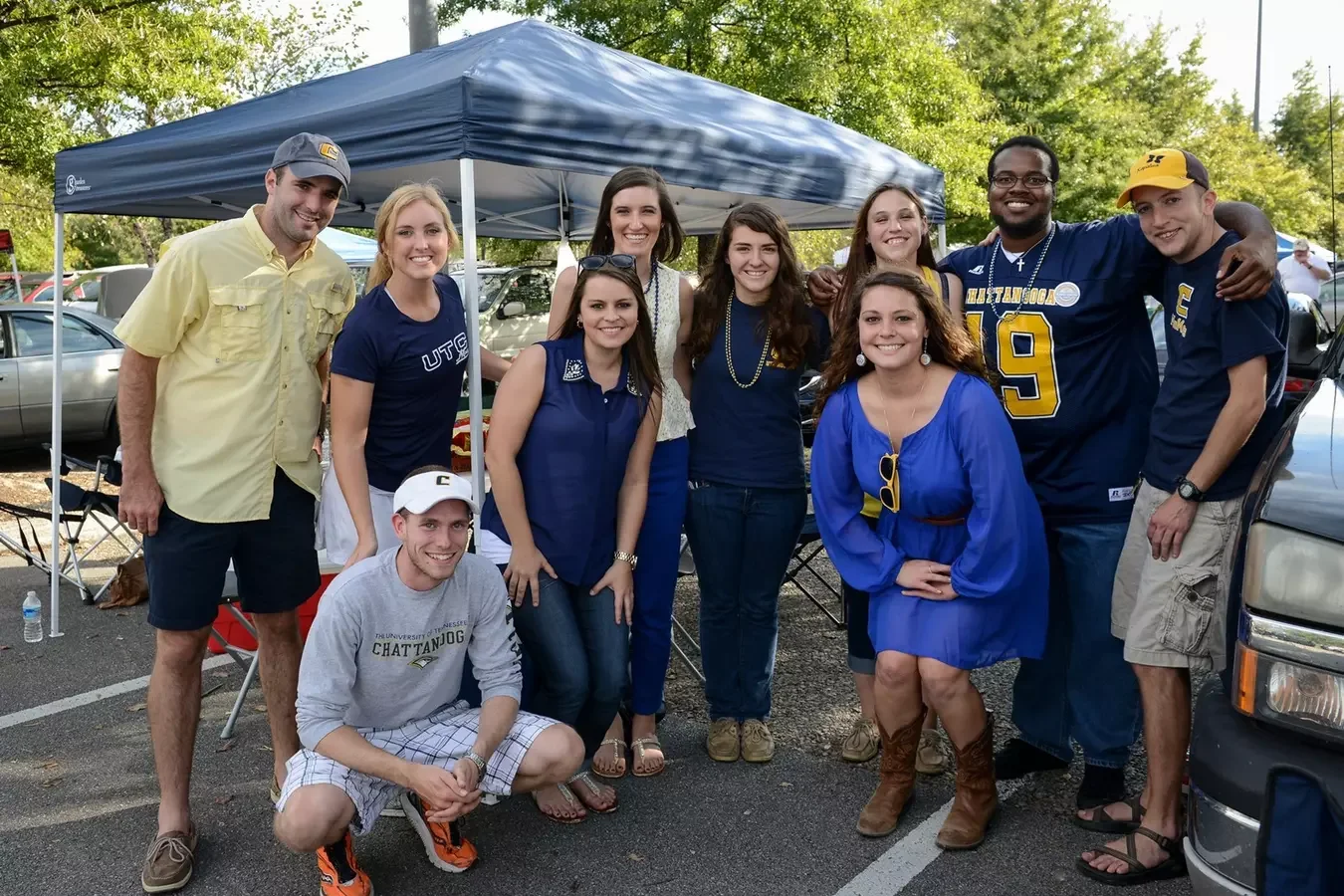 Group of young adults, some wearing University of Tennessee Chattanooga gear, gathered outdoors under a canopy tent at a tailgate party, smiling for a photo.