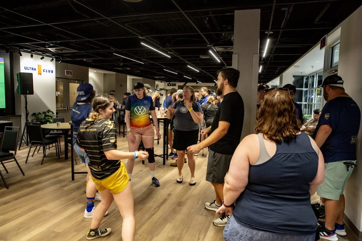 Group of people dancing and socializing in a room with wooden floors and modern lighting, some wearing sports jerseys and colorful clothing.