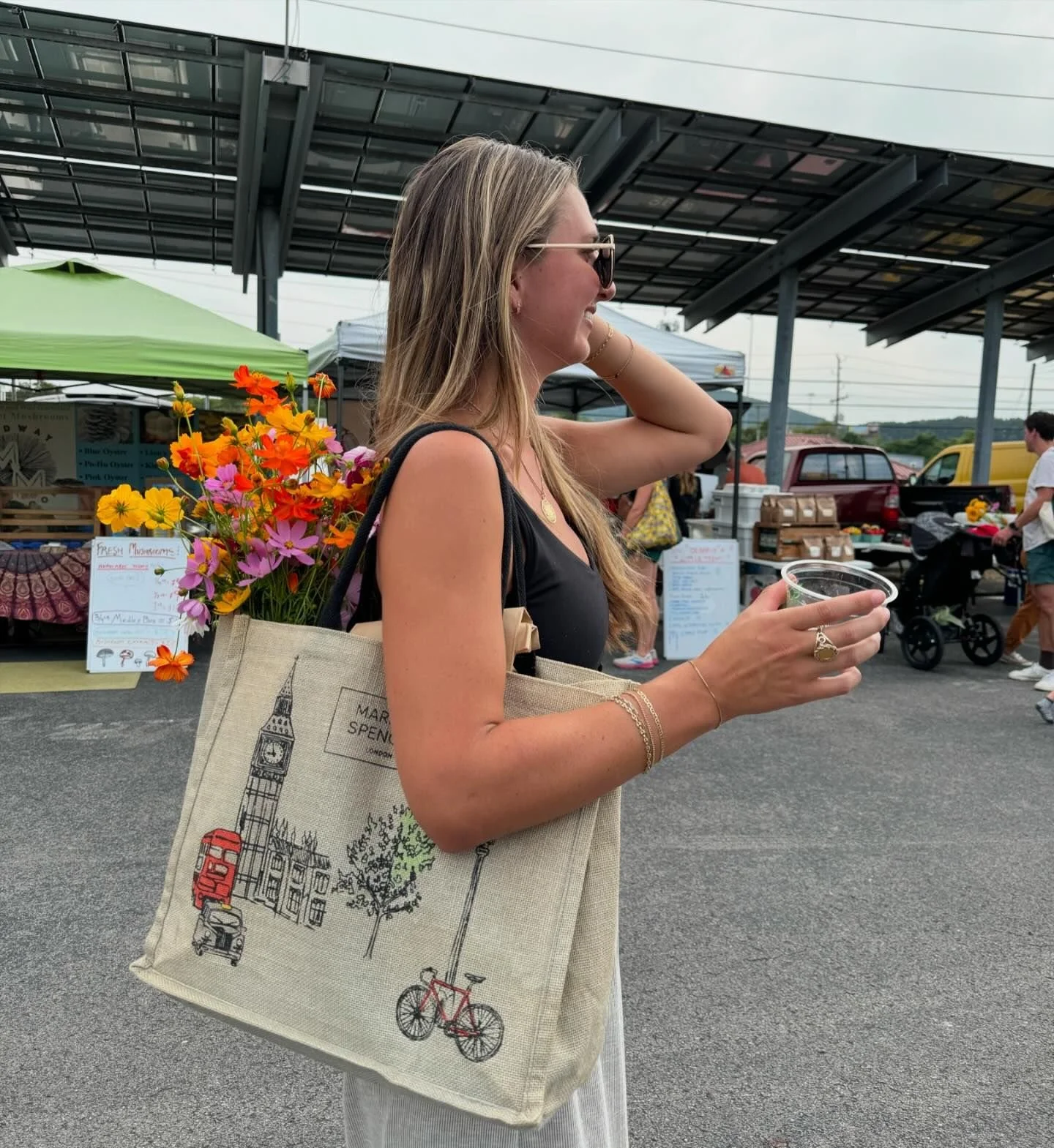 A woman with long blonde hair, wearing sunglasses and jewelry, holding a cup of beverage and a beige tote bag with a London-themed illustration, standing at an outdoor market. The market has stalls under tents, with people browsing, and a large solar panel structure overhead.