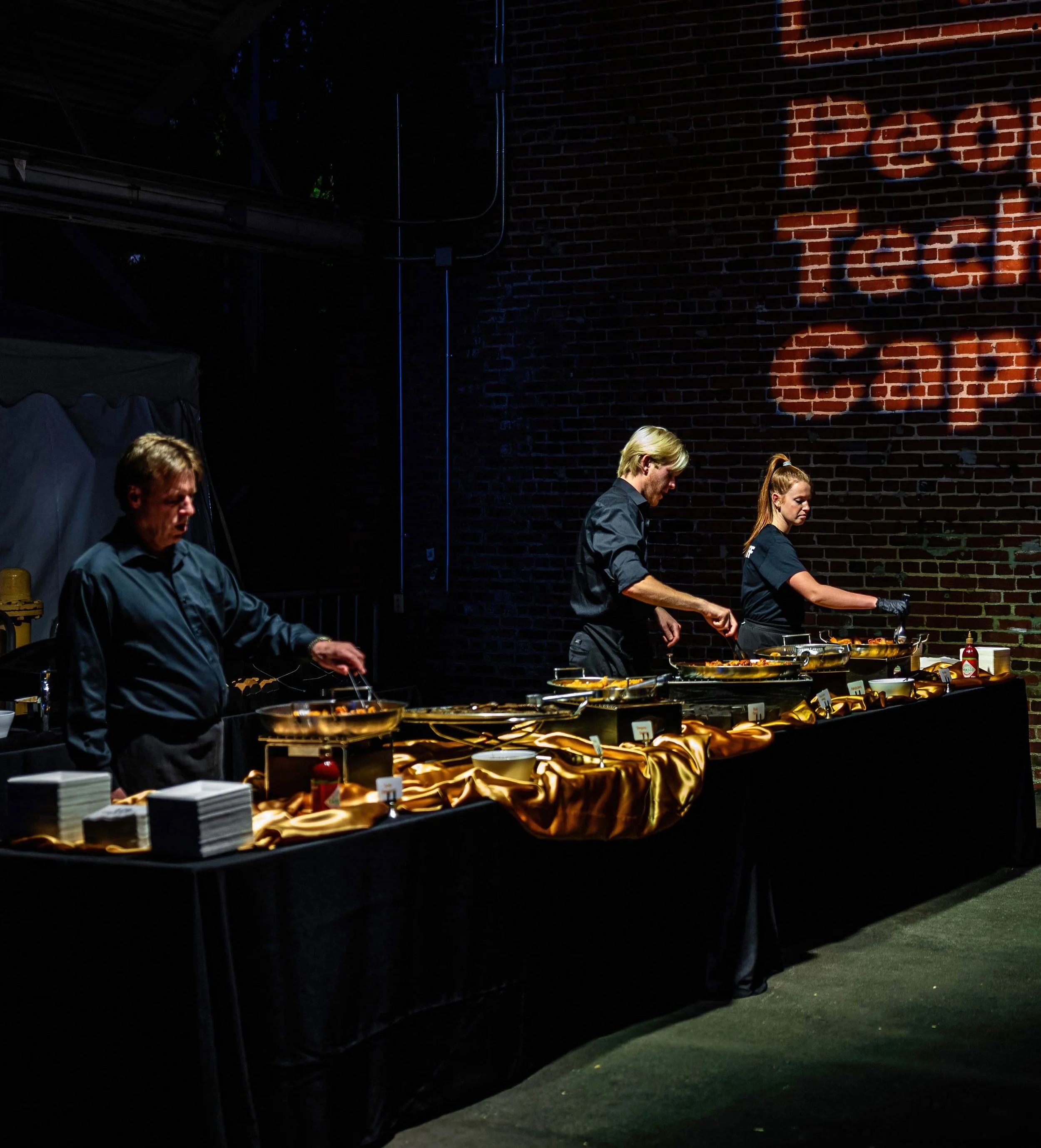 A buffet table with food trays and plates, set against a dark brick wall with a light projection of the word 'BASH' on it, in an outdoor or semi-outdoor setting.