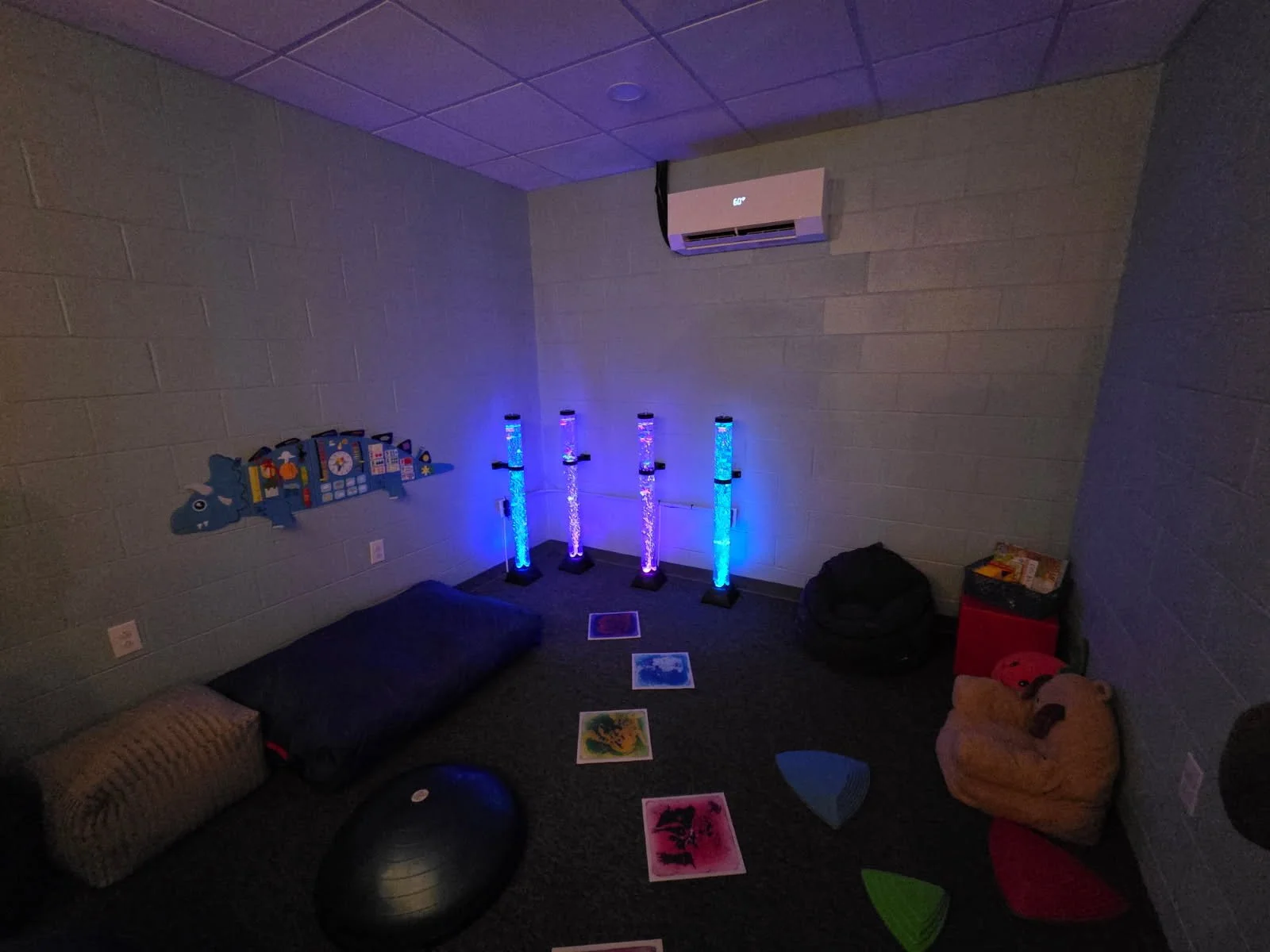A cozy playroom with gray brick walls and a gray carpeted floor. Brightly lit bubble tubes with colorful lights are in the corner. The walls have some playful decorations, and there are various toys including a large teddy bear, a black exercise ball, and some colorful mats or pillows scattered on the floor.