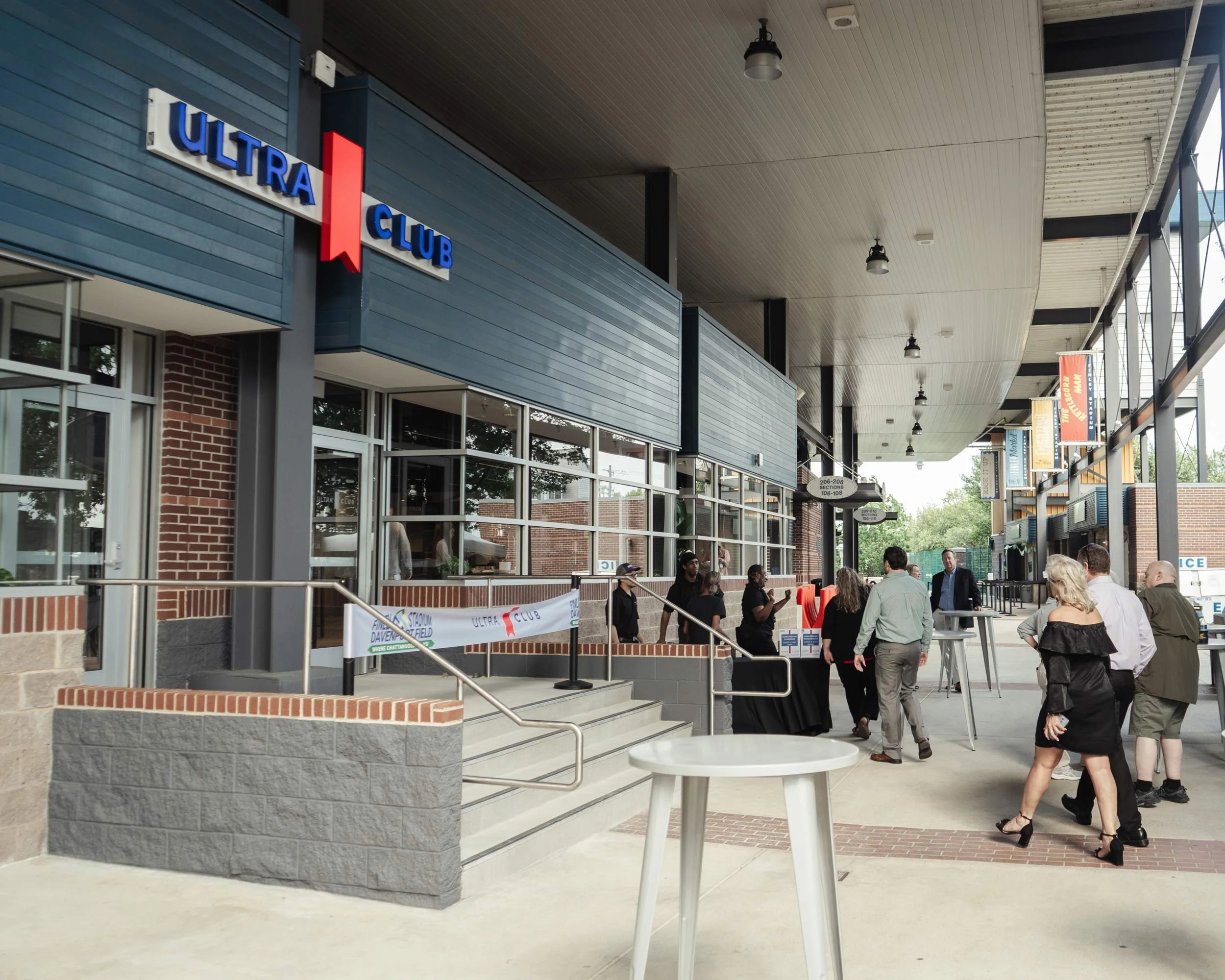 Exterior view of a stadium entrance with a sign reading 'ULTRA Club'. There are people walking and standing outside, and the area has a modern design with metal, brick, and glass elements. Banners and signs can be seen along the walkway.