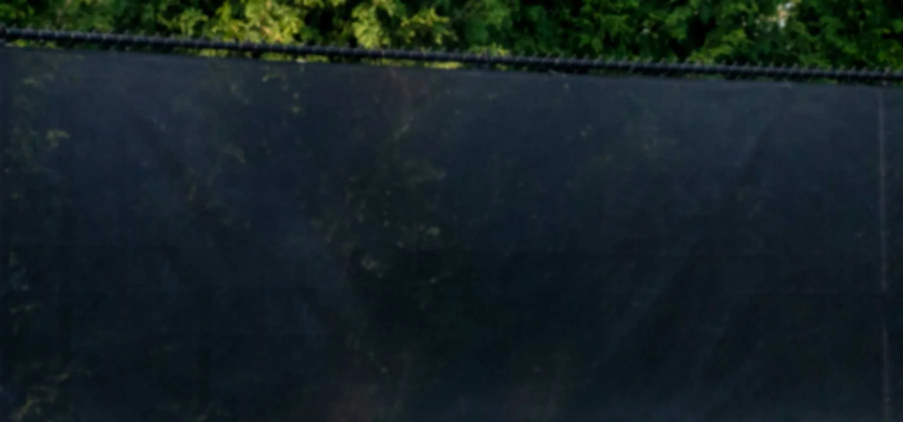 A dark, shiny surface reflecting trees and sky, with a black metal pipe or beam running horizontally across the upper part of the image. There is greenery visible behind the surface.