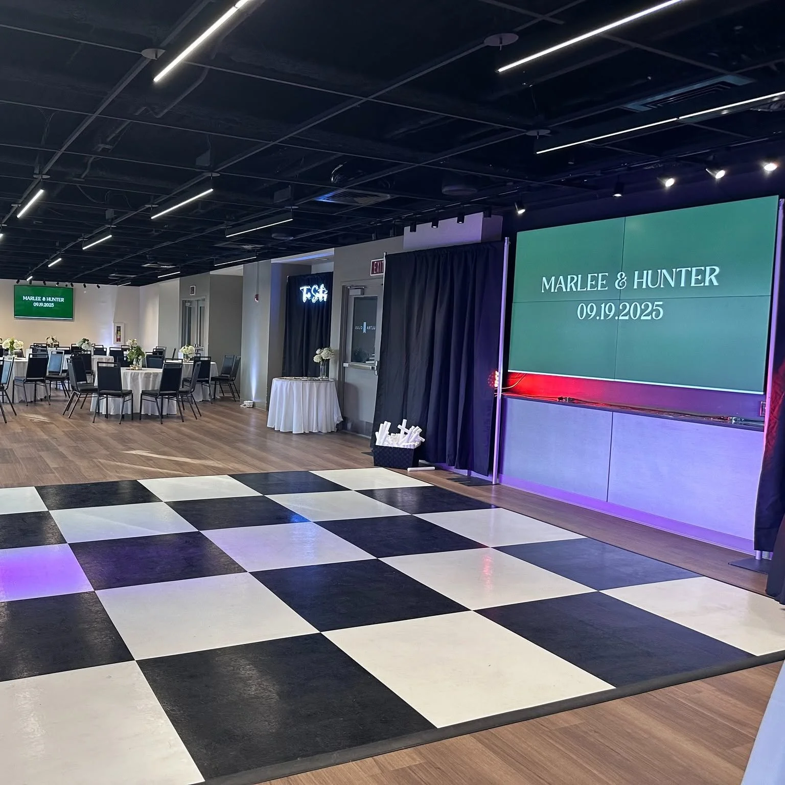 Event space with black and white checkered dance floor, round tables with white tablecloths and flower centerpieces, large screen displaying names 'Marlee & Hunter' and date '09.19.2025', and black curtains with some floral arrangements.