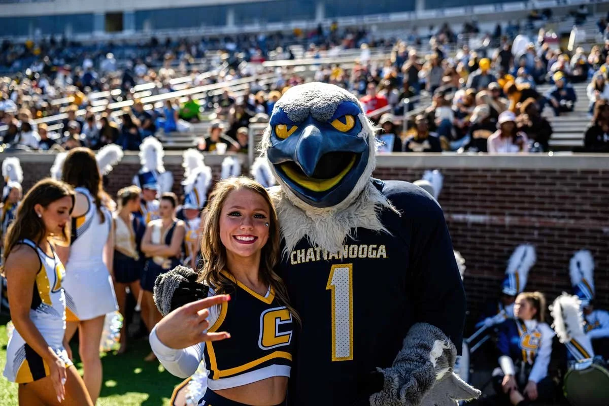 A woman and a bird mascot in sports uniforms at a football game, with cheerleaders and spectators in the background.