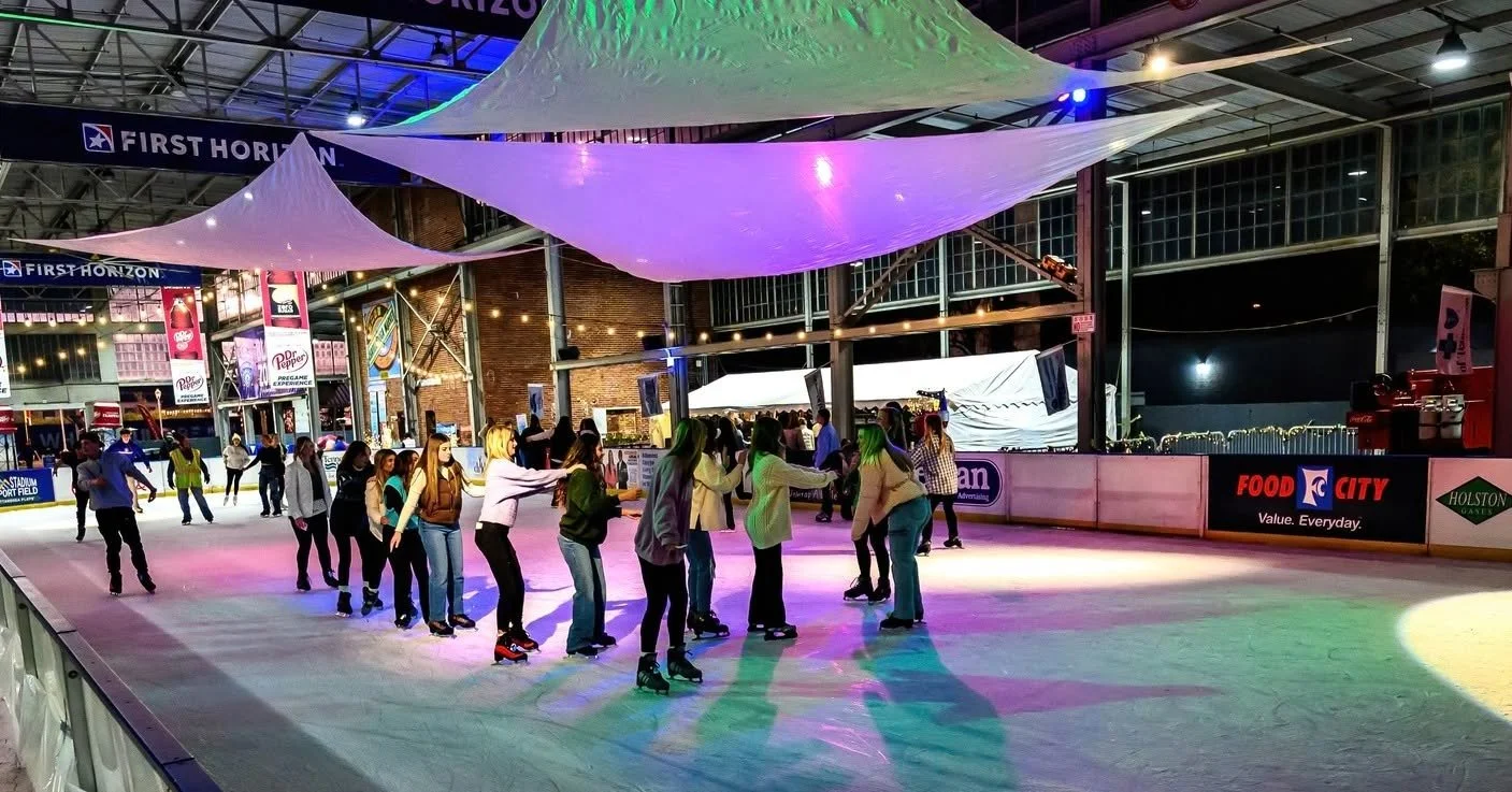People skating on an indoor ice rink at night. There are colorful lights and hanging fabric decorations above. Commercial signs and a brick wall are visible in the background.