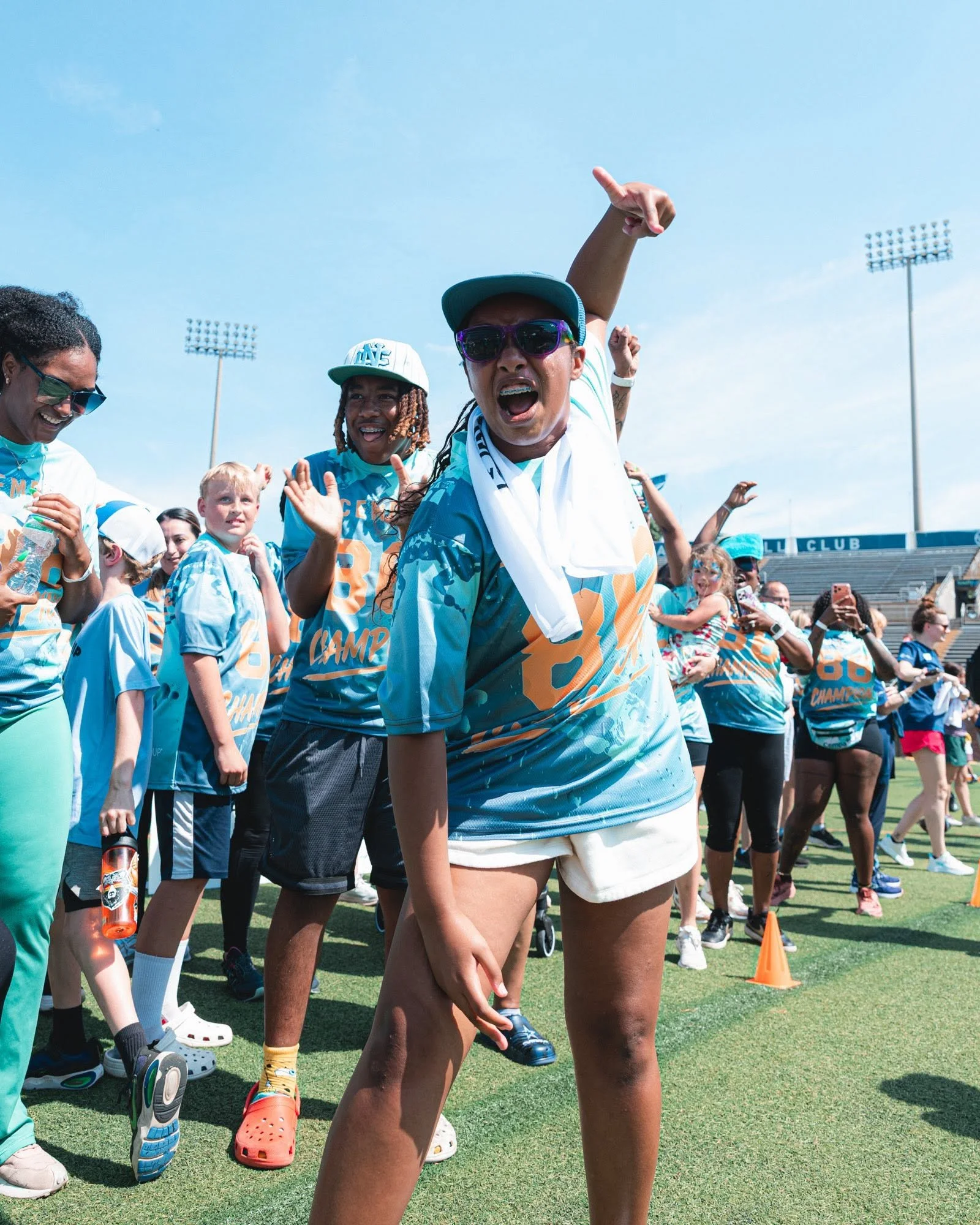 Group of people gathered outdoors on a sports field, some wearing bright blue shirts and caps, with one person in the center making a gesture with their hand, crowds and stadium lights visible in the background.