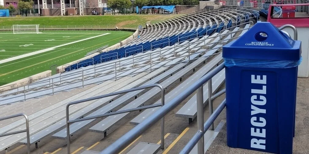 Empty bleacher seats at a sports field with a blue recycling bin labeled "RECYCL E" in the foreground.