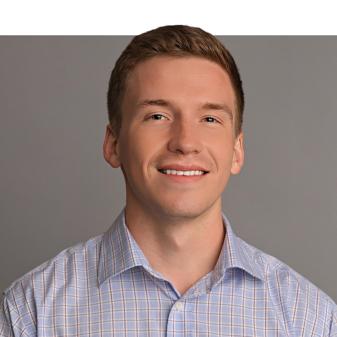Portrait of a young man with short light brown hair, wearing a plaid button-down shirt, smiling against a plain gray background.