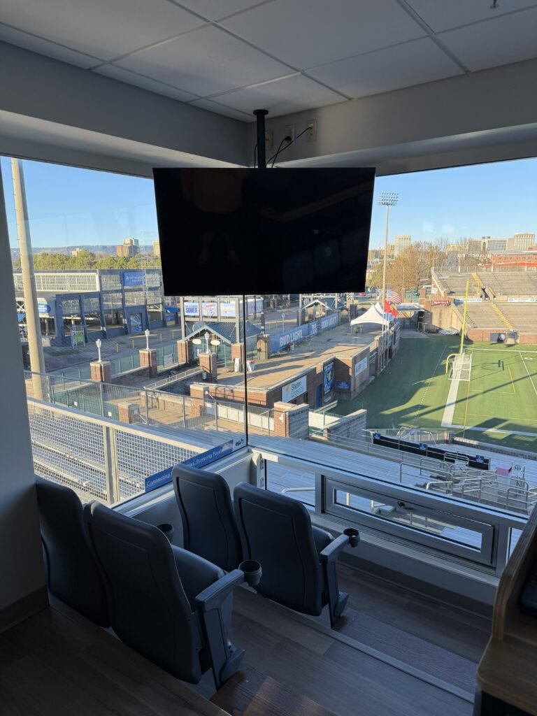 View from a media box at a sports stadium, showing the empty seats and field outside with a large television screen hanging from the ceiling in the interior.
