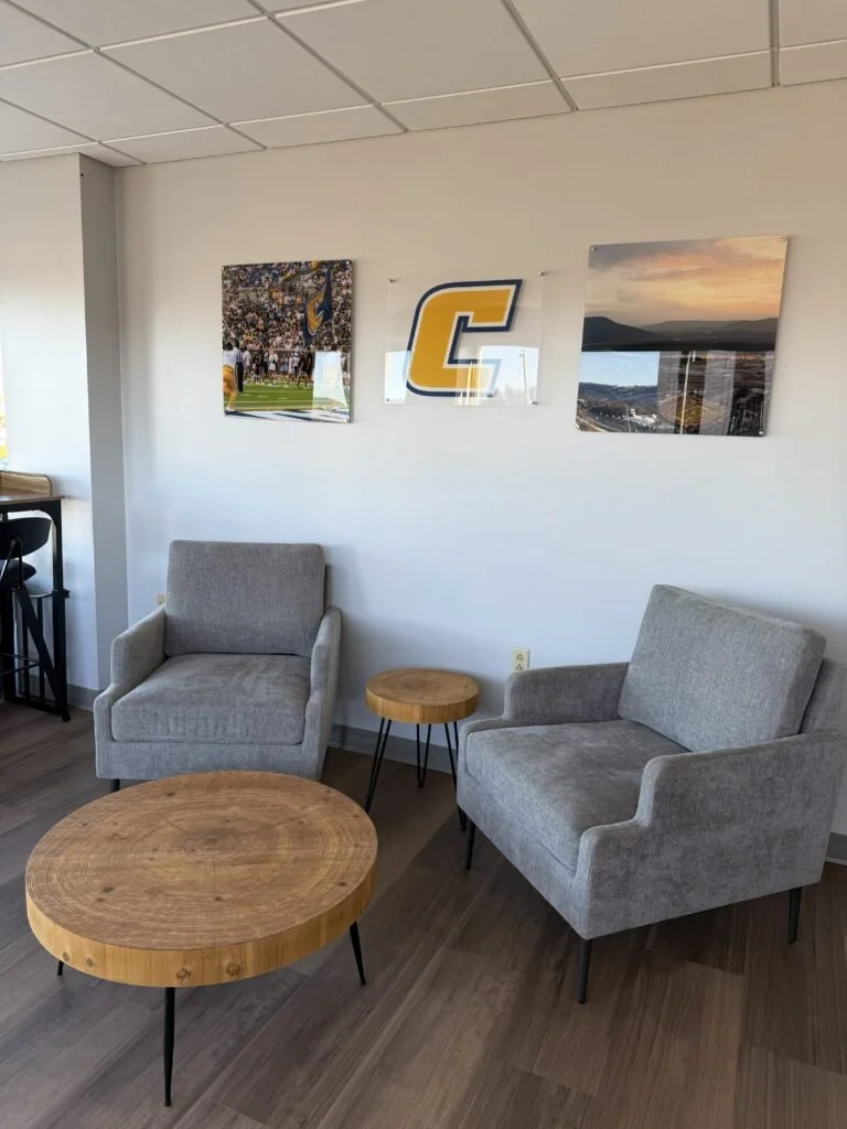 A cozy sitting area with two light gray armchairs, a large wooden coffee table, and a smaller wooden side table placed between the chairs, with framed sports and landscape photos on the white wall behind them.