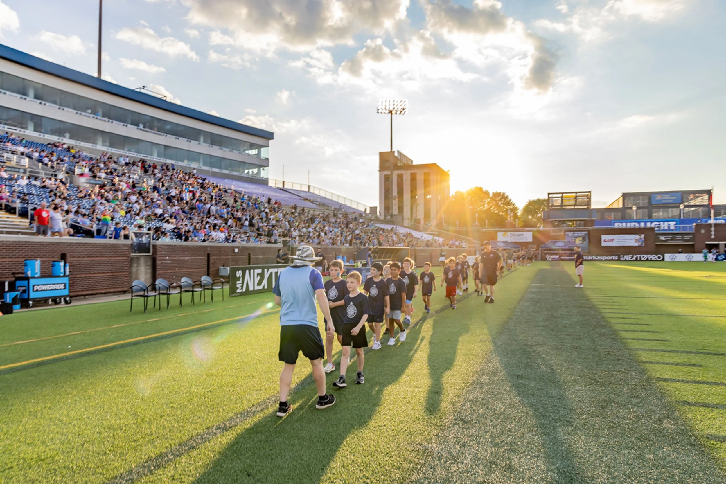 Children walking in a line on a sports field during a sports event with a packed stadium in the background, under a partly cloudy sky with sunlight.