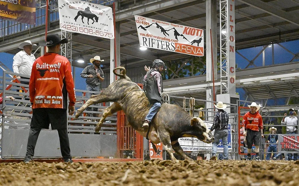 Rodeo event with a rider on bucking bull and several cowboys in Western hats and cowboy attire, in an arena with banners overhead.