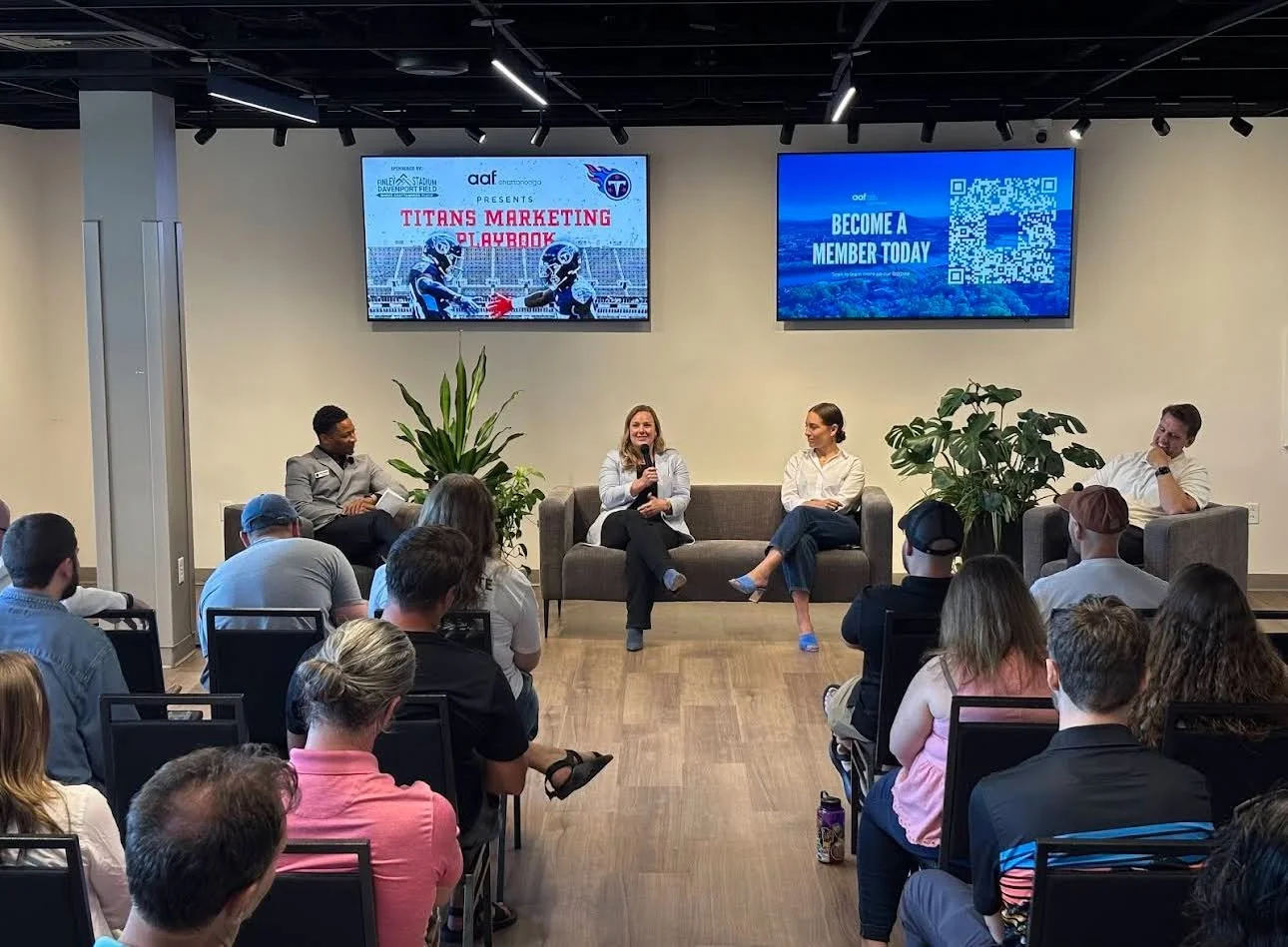 A panel discussion at a Titans marketing event with four speakers on a stage, two women and two men, seated on a sofa and chairs. Audience members are seated facing the panel, with some taking notes or listening attentively. Two large screens behind 