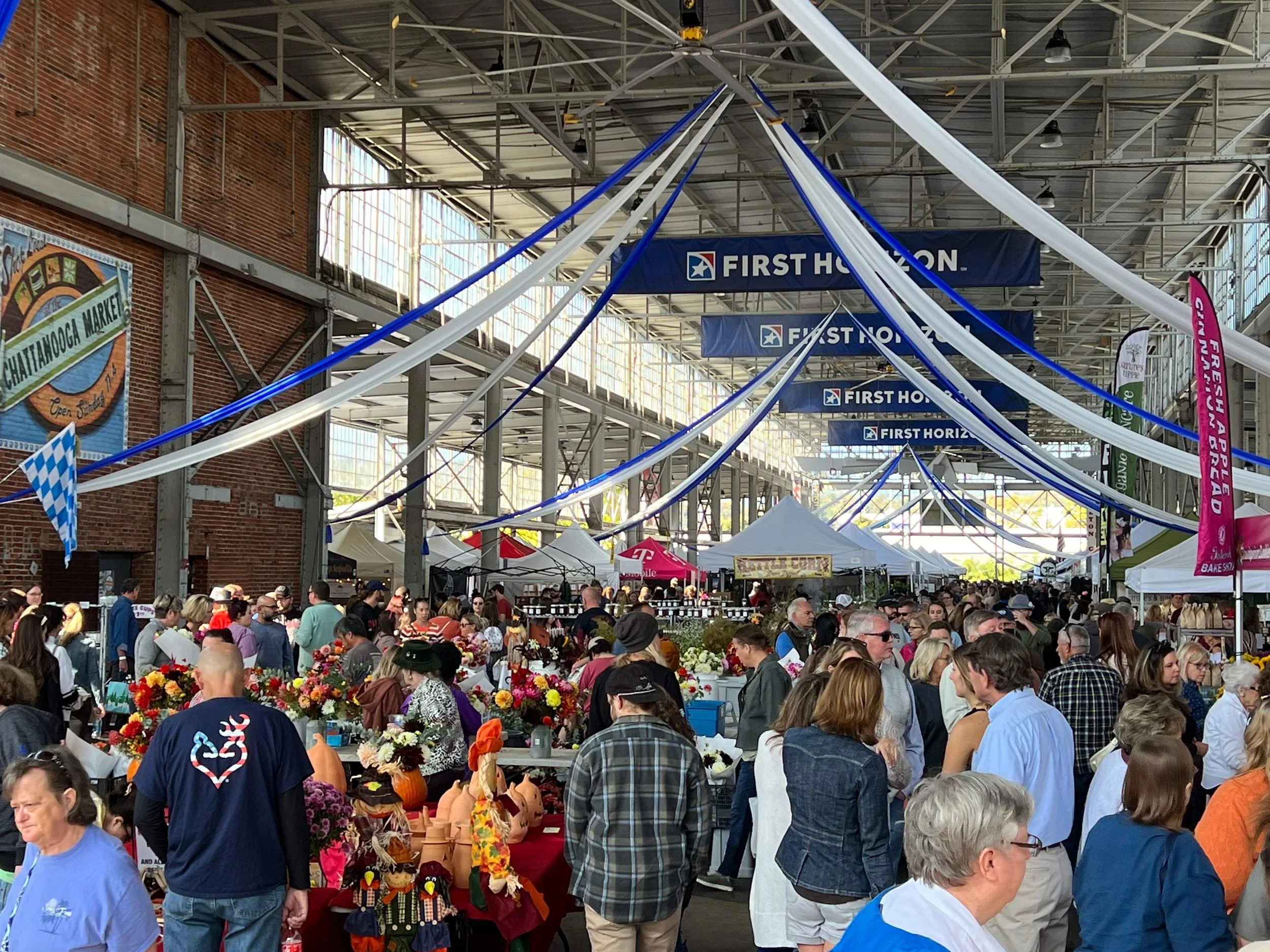 Crowd of people at an indoor market or festival with vendors and various booths, some displaying flowers and crafts, under a high metal roof with banners reading "First Horizon".