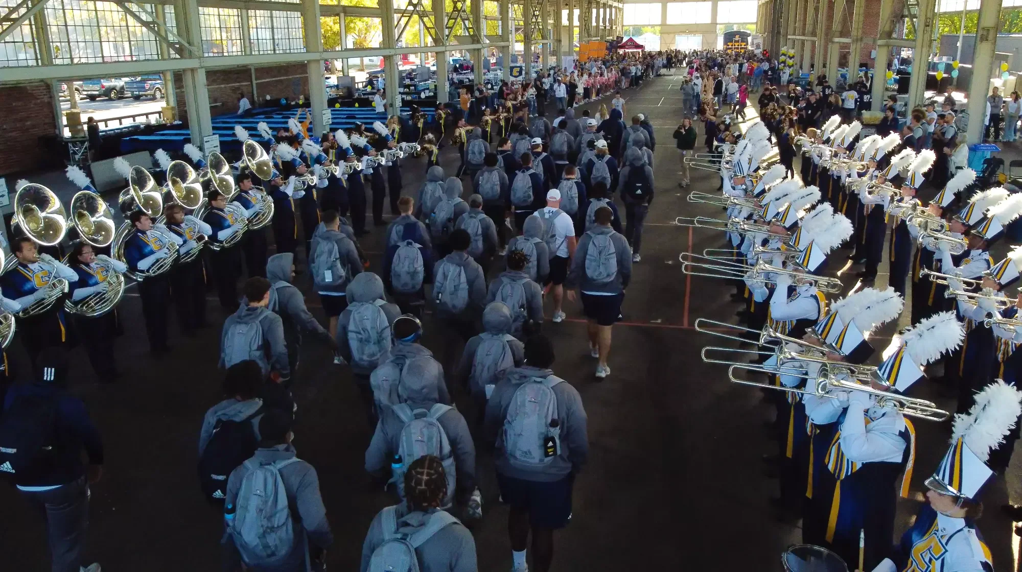 A large indoor area with a marching band lined up on one side and a crowd of people walking through. The band is dressed in uniforms with hats adorned with white feathers, playing brass instruments like tubas and trumpets. The crowd appears to be stu