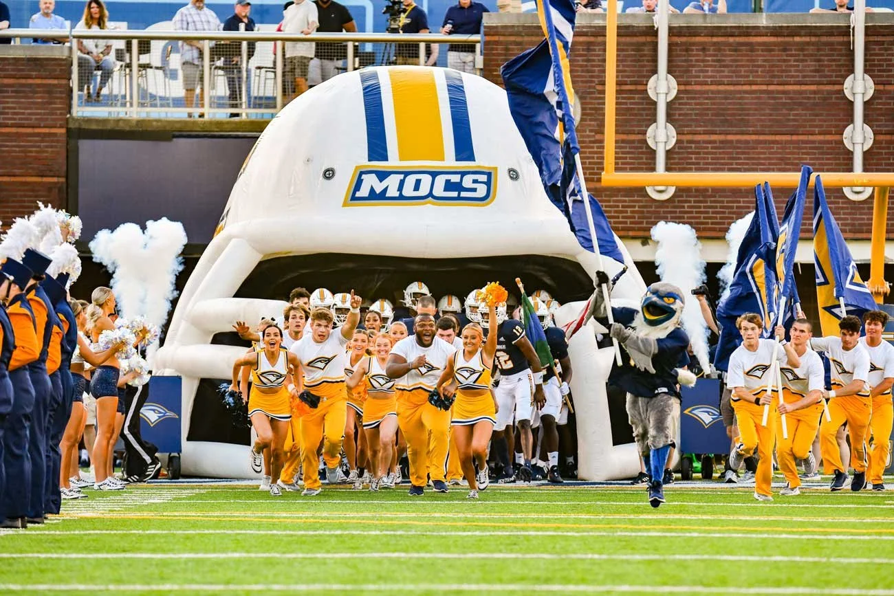A high school football team and cheerleaders running onto the field through a mascot tunnel, with fans and cheerleaders on the sidelines celebrating.
