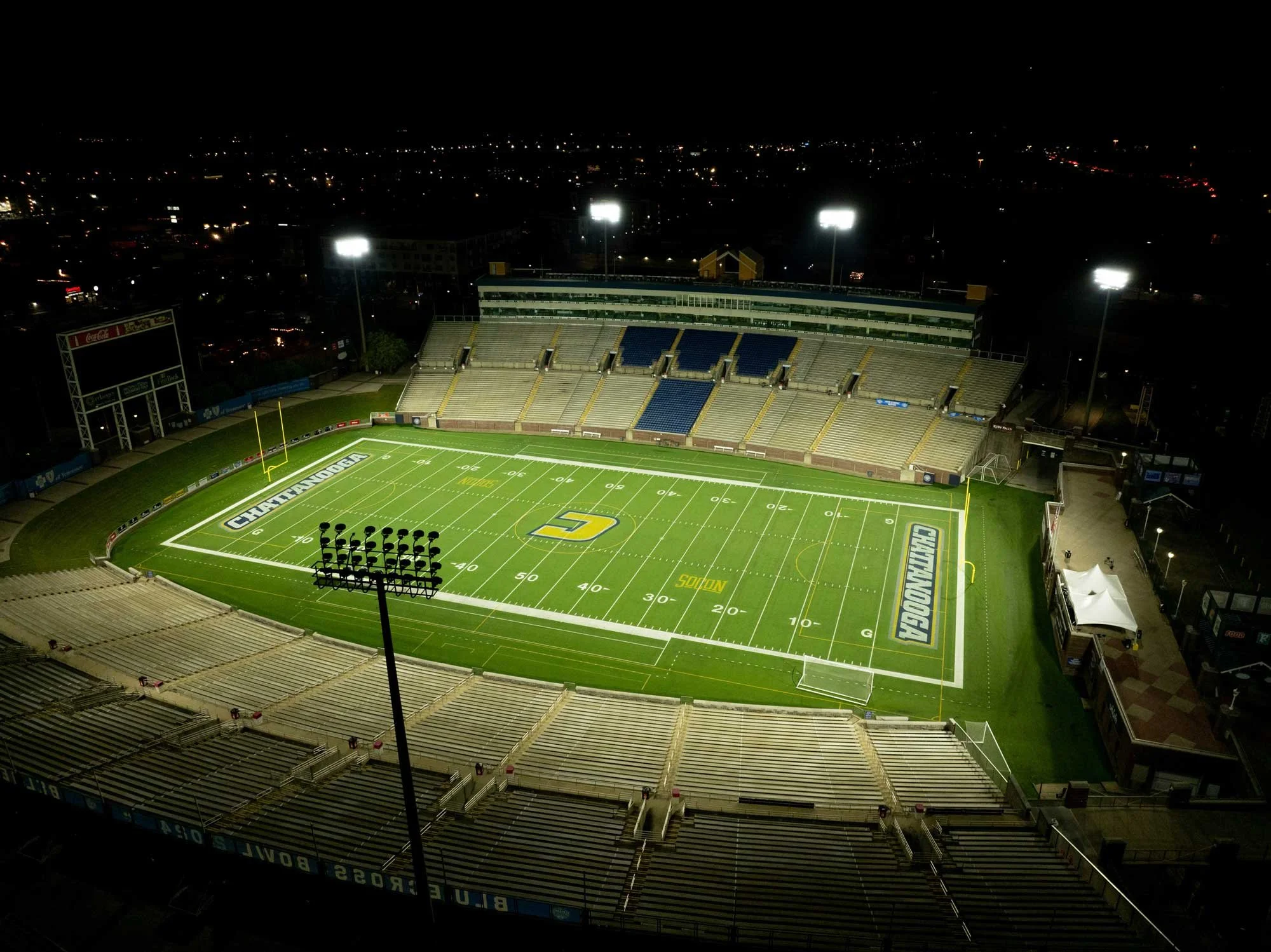 Empty football stadium at night with bright floodlights illuminating the field. The stadium has empty seats in yellow and blue, with the field marked for a game and the word 'Reseda' visible on the turf near the end zone. City lights can be seen in the background under a dark sky.
