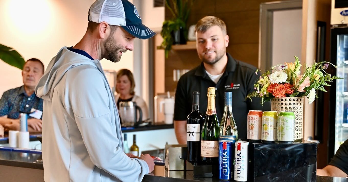 A man in a hoodie and baseball cap buying drinks at a bar counter, with a bartender and customers in the background and bottles, cans, and a vase of flowers on the counter.