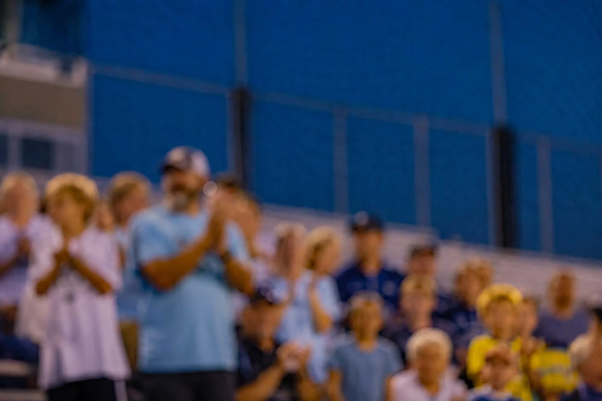 A group of people, including children, sitting and standing at an outdoor event, possibly a sports game or concert, with a large blue wall or structure in the background.