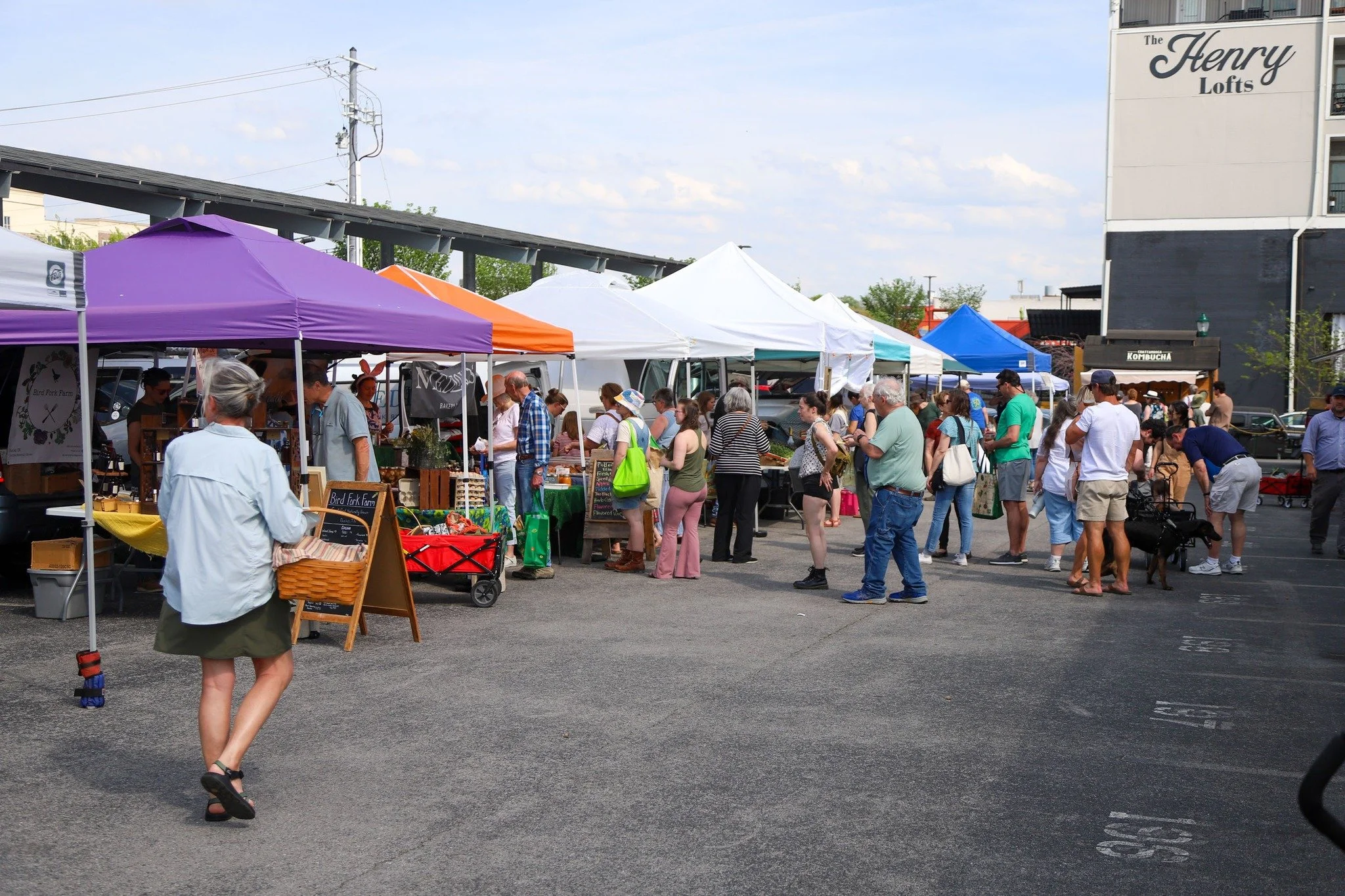 People walking and shopping at outdoor market with vendor tents and store building in background.