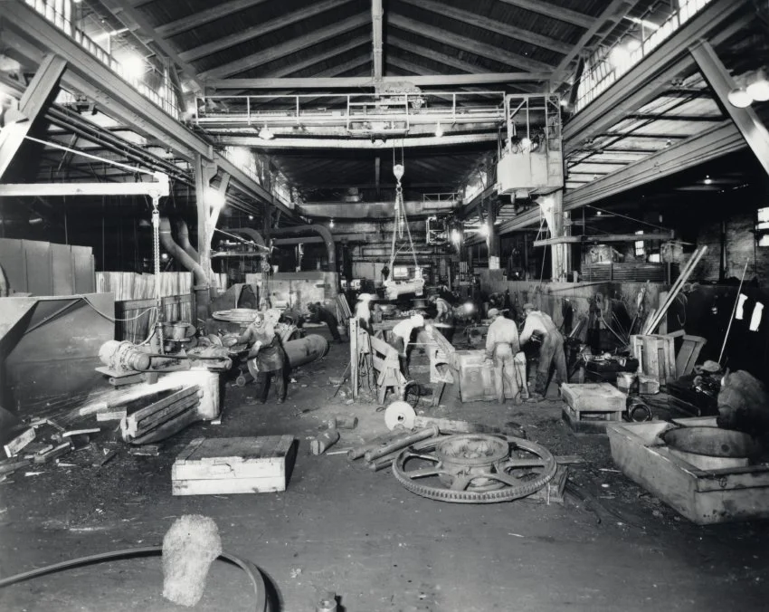 Black and white photo of an industrial workshop with workers engaged in manufacturing or welding, surrounded by machinery, tools, and equipment.