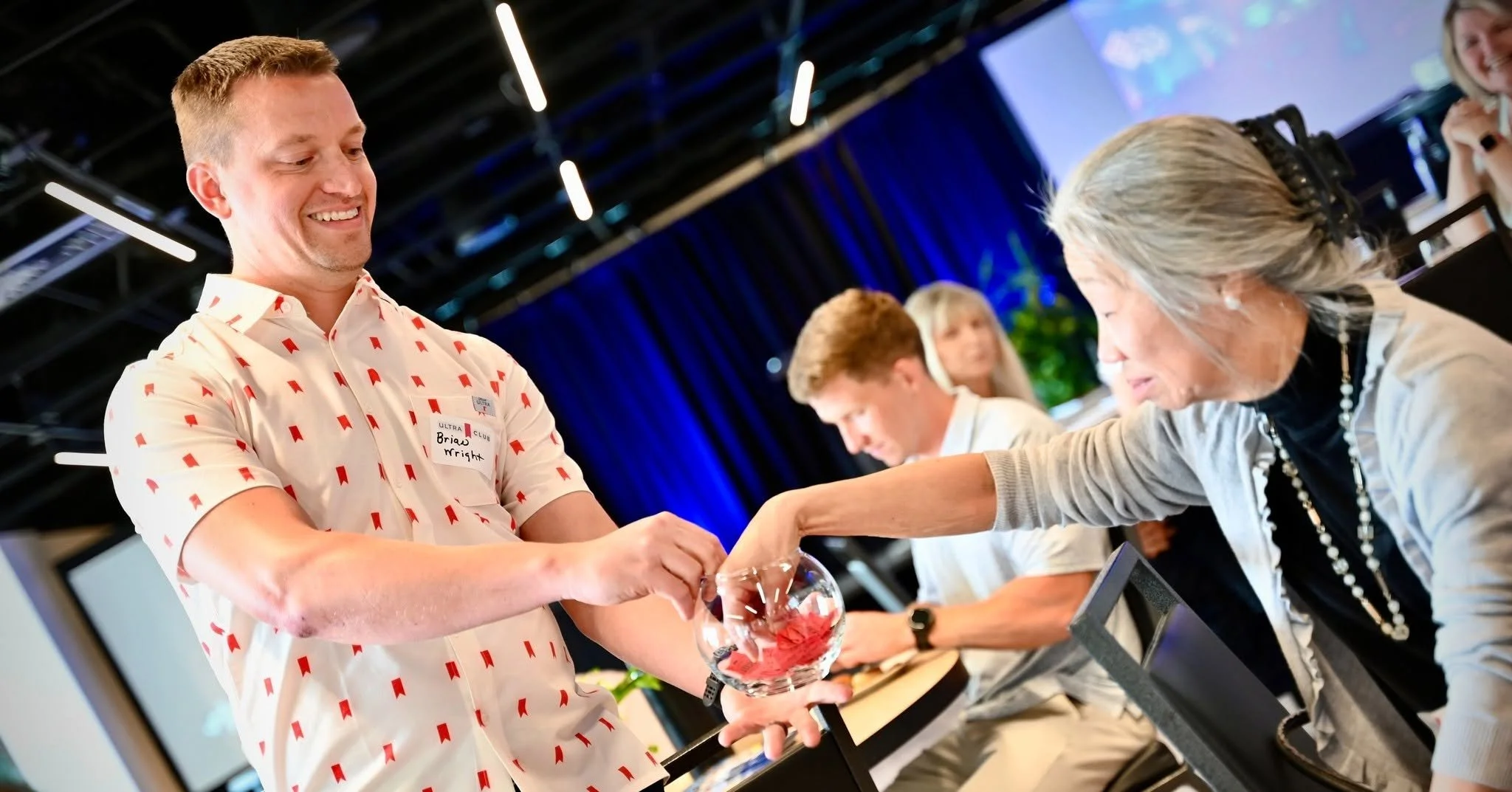 A smiling man in a white shirt with red arrow patterns is shaking hands with an older woman who is smiling and leaning forward. They are at a table with a glass bowl in front of the man. Other people are in the background, and the setting appears to 
