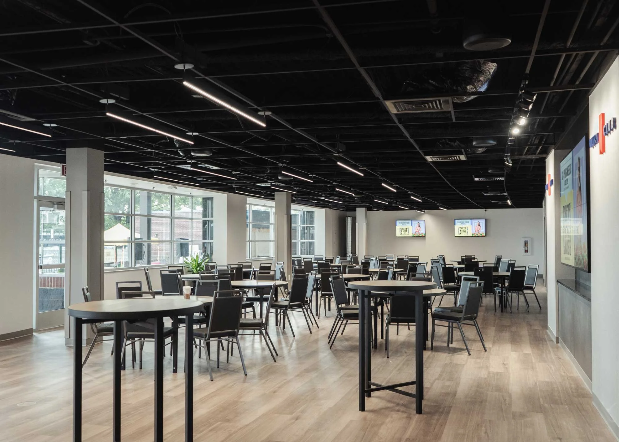 Empty indoor space with tables and chairs, large windows allowing natural light, modern ceiling with linear lighting, digital screens on the walls.