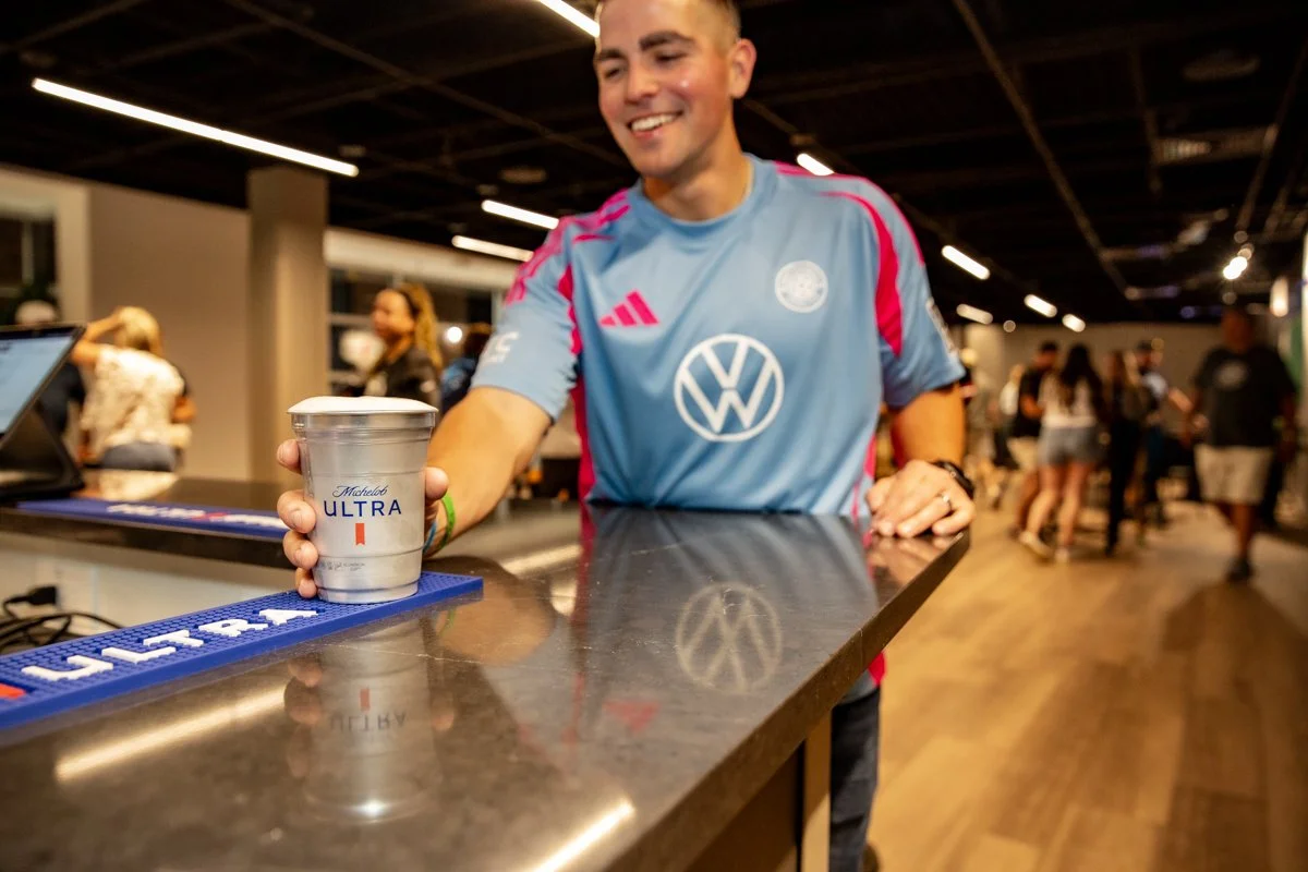 A man in a blue sports jersey smiling and handing a silver cup labeled 'Michelob Ultra' to someone at a bar or counter.