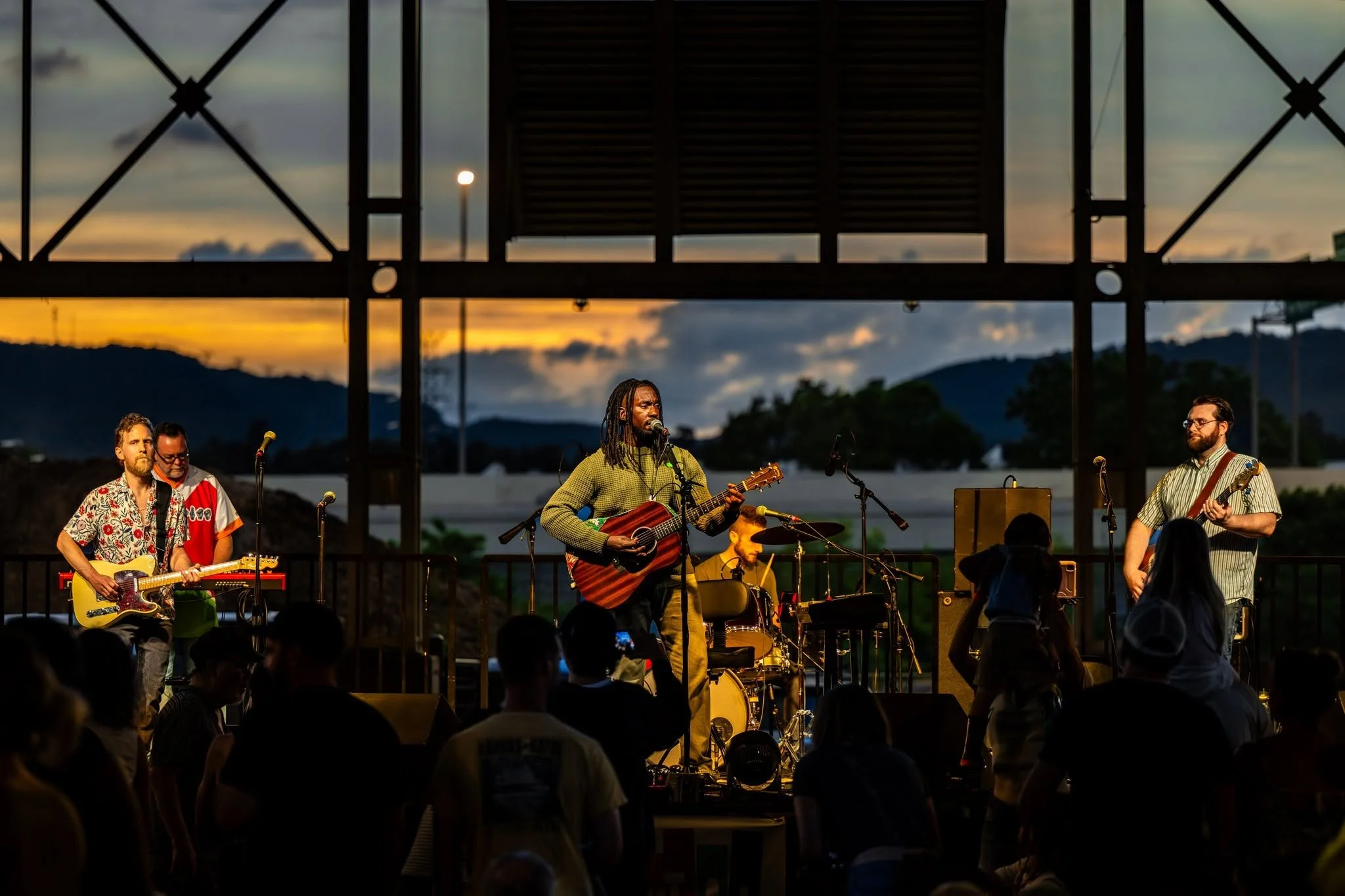 A band performing on an outdoor stage at sunset, with mountains and cloudy sky in the background. The band includes a man with dreadlocks singing and playing a guitar, and other musicians playing guitars and drums. An audience is watching.