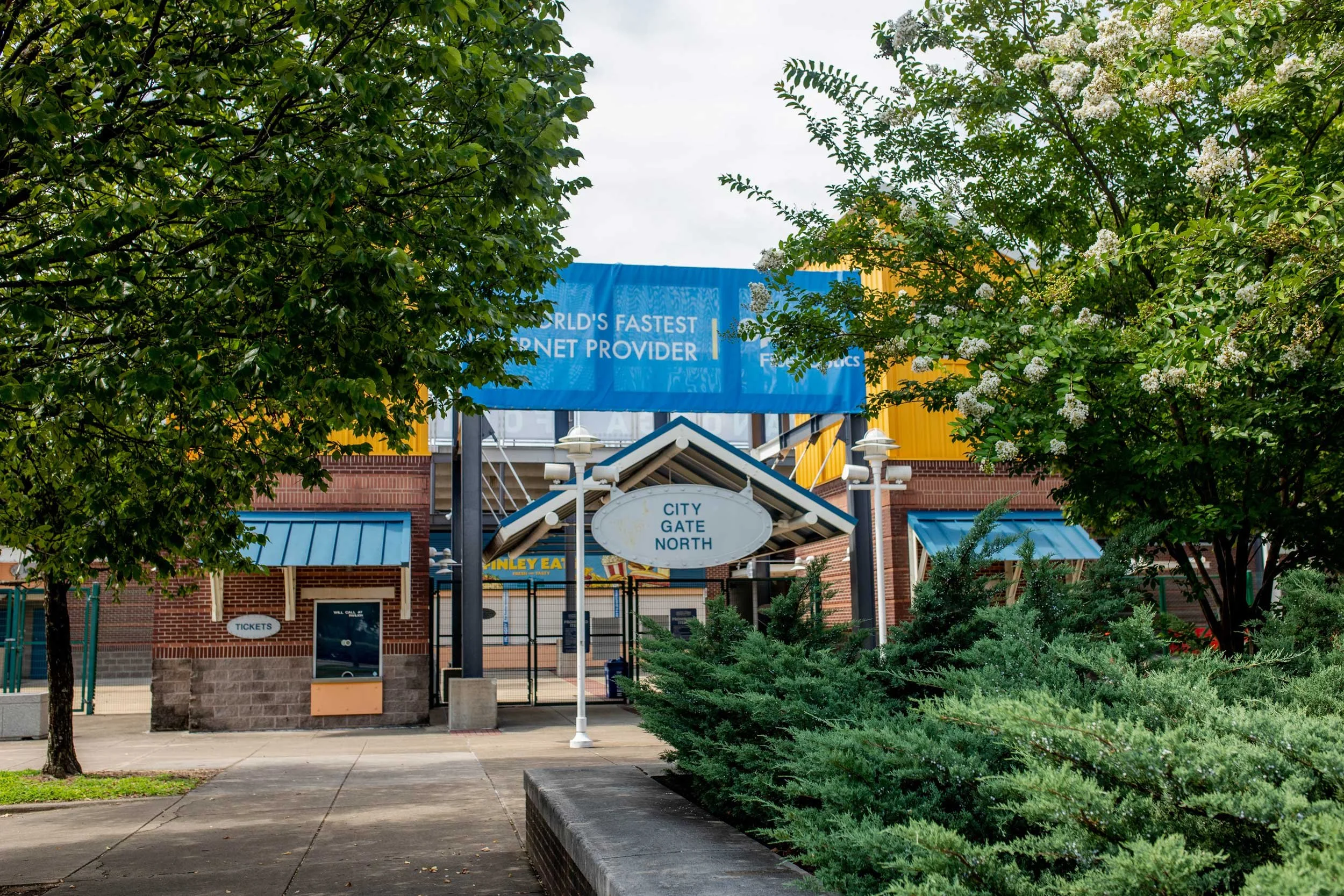 Entrance to a stadium with signs displaying 'City Gate North' and 'Tickets,' surrounded by trees and greenery.