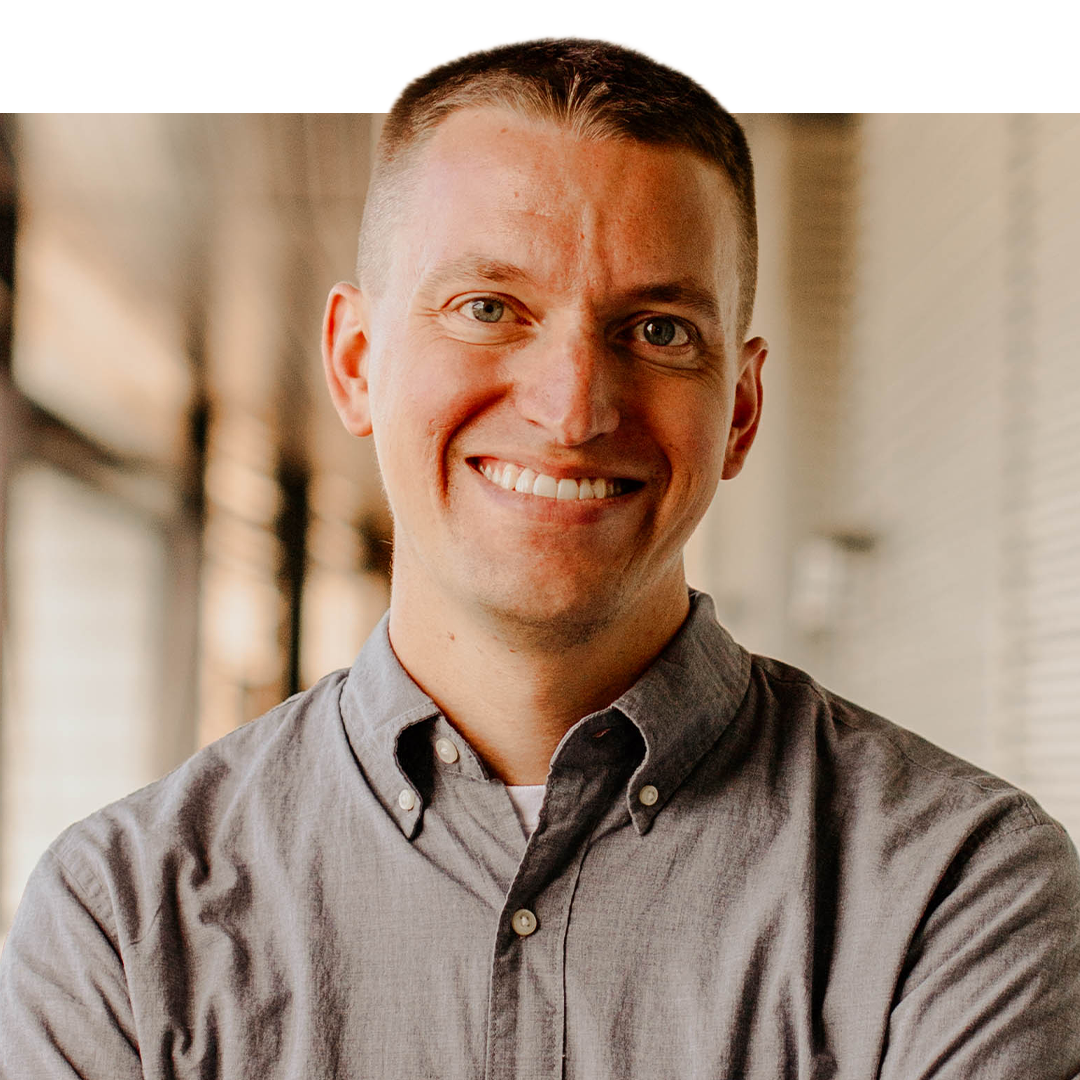 A smiling man with short hair, wearing a gray button-up shirt, standing in a well-lit indoor space with blurred background.