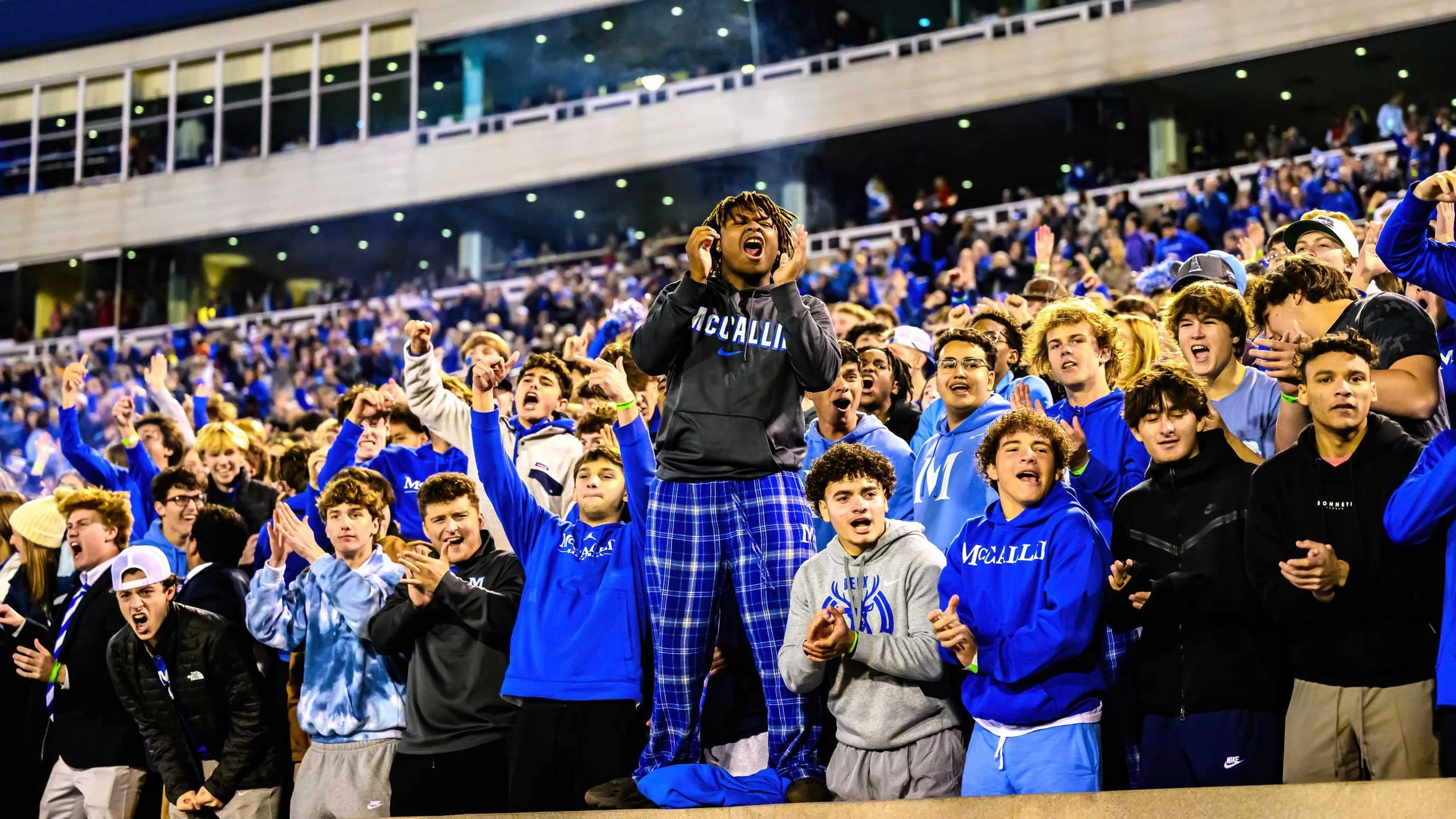 Group of young people cheering and celebrating at a stadium, some wearing blue hoodies and jackets, and one person in the center standing on a ledge with their hands on their ears.