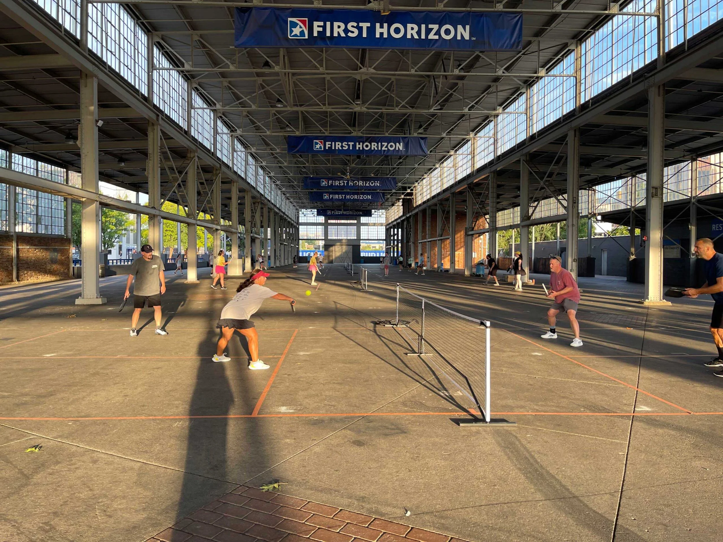 Indoor court with a sign that says 'First Horizon'. Several people are playing, some are in yellow outfits, and others are in darker clothing. The court is divided by netting, and the structure features large glass windows letting in natural light.
