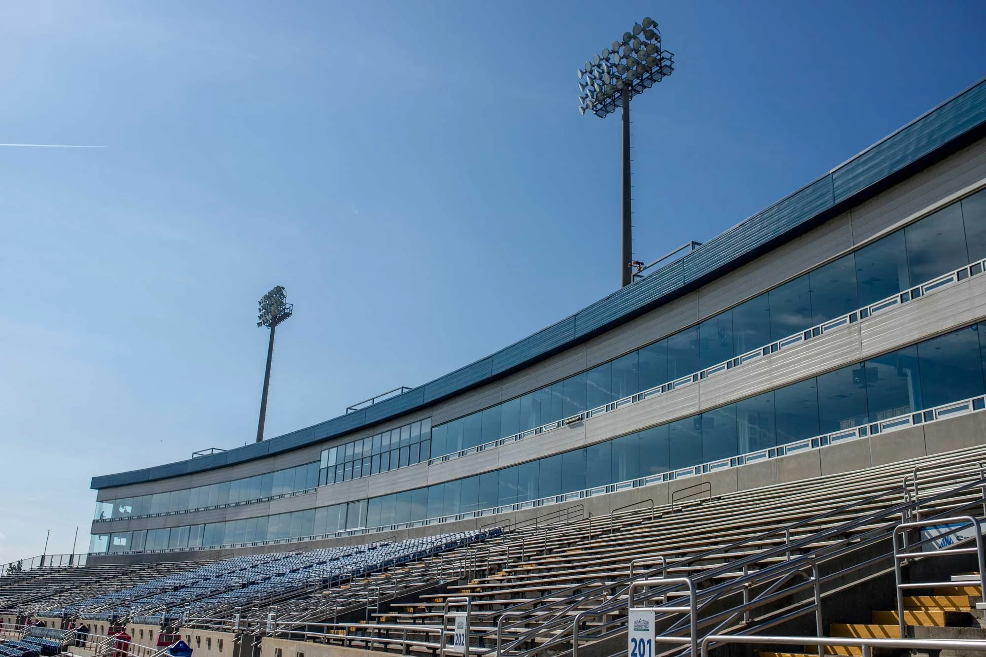 Empty outdoor stadium seating with a modern glass-fronted building in the background, two tall floodlights, and a clear blue sky.