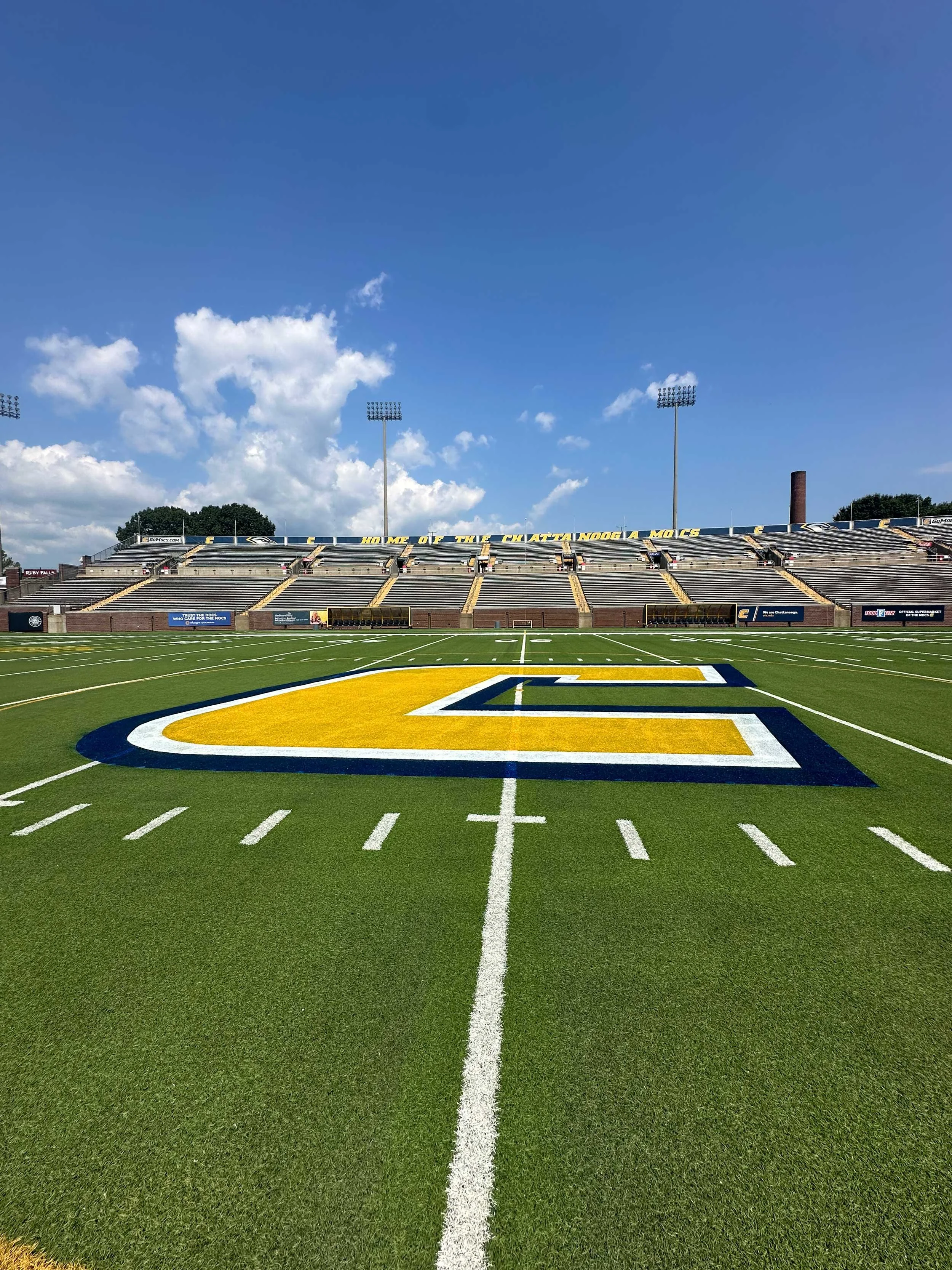 A football field with yard lines, a large logo at the center, and empty bleachers in the background under a clear blue sky.