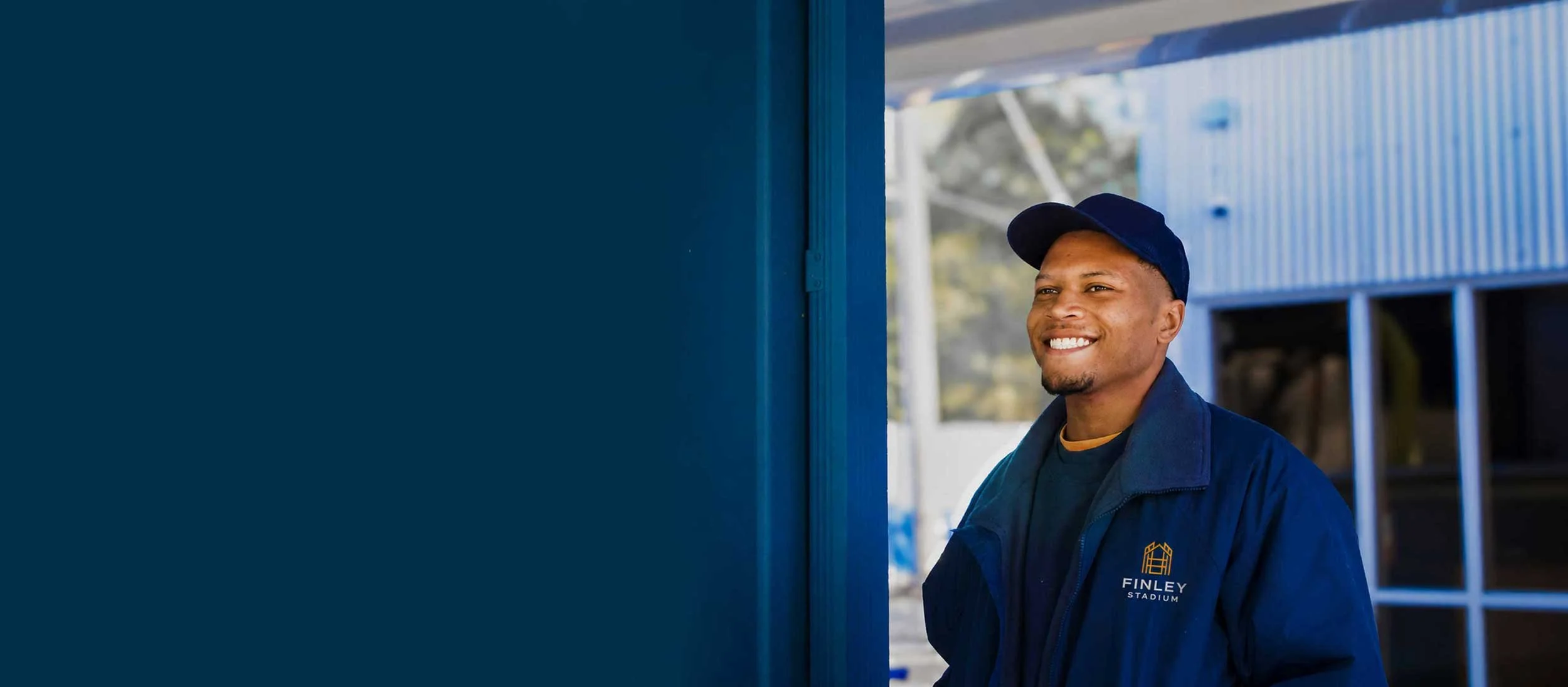 A smiling man wearing a navy blue cap and jacket with a logo reading 'Finley Stadium' stands outside near a blue building.