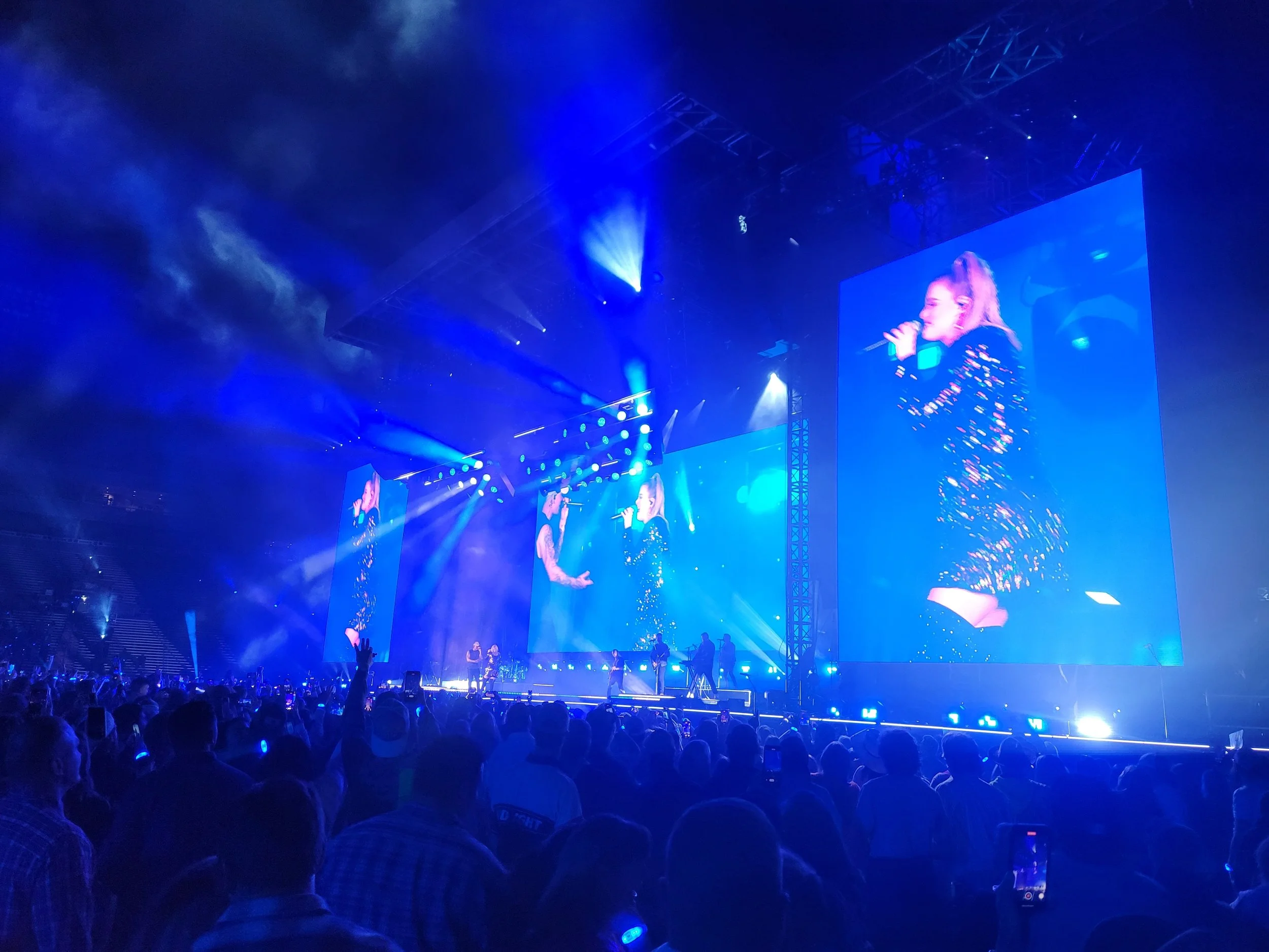 A concert stage illuminated with blue lights and fog, showing a large screen with an image of a woman singing into a microphone, surrounded by an audience.