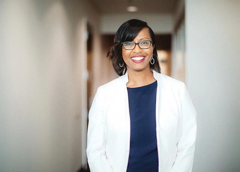 A professional woman with glasses and earrings, wearing a white blazer and navy blue top, smiling in a well-lit hallway.