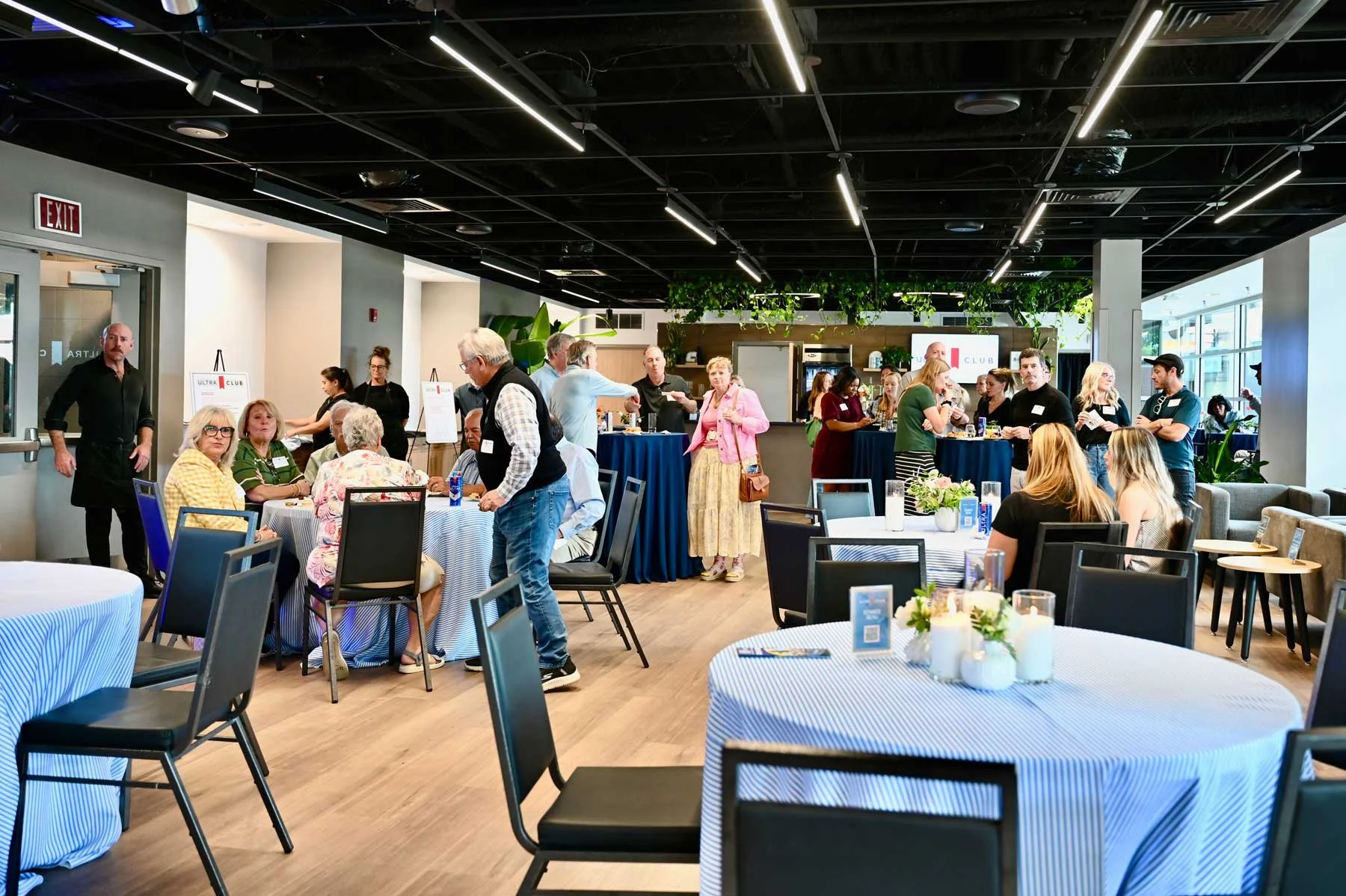 Interior of a modern event space with people socializing, eating, and talking. Round tables with blue and white striped tablecloths, some with floral centerpieces. Several standing guests and tables with drinks and food, set in a bright room with large windows, greenery, and a black ceiling with striped lighting.