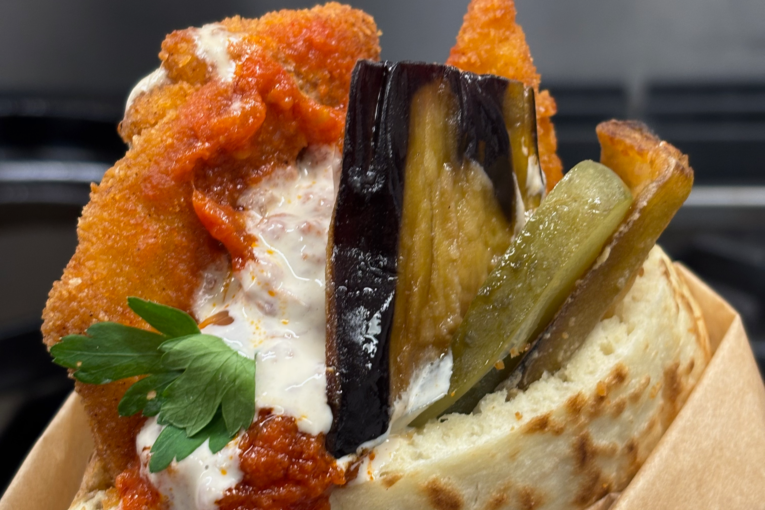 A close-up of a slice of bread topped with fried chicken, tomato sauce, melted cheese, and pickles, garnished with a parsley leaf.