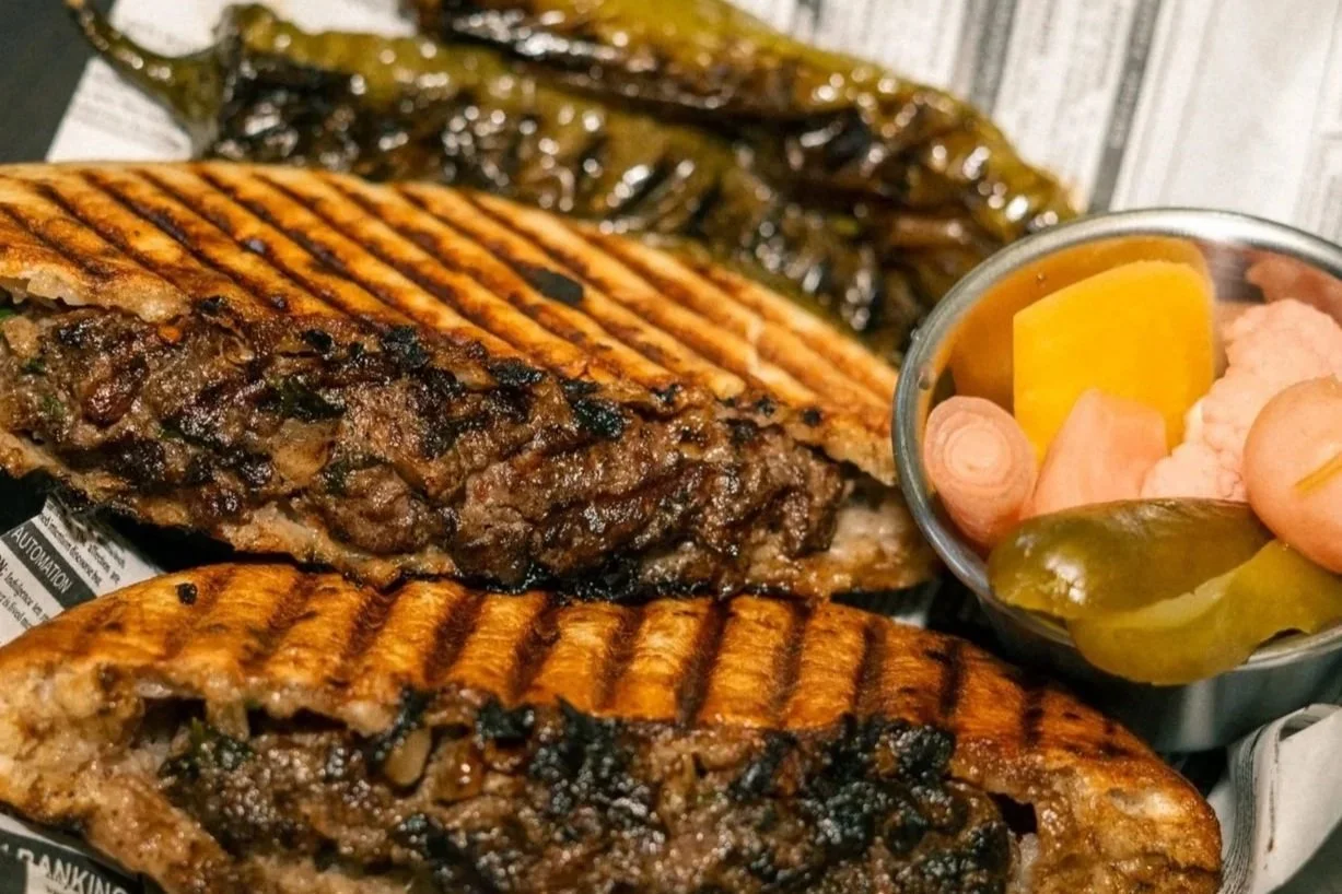 Two grilled steak sandwiches with visible stuffing of cooked vegetables, served with pickles and a side of grilled banana peppers.
