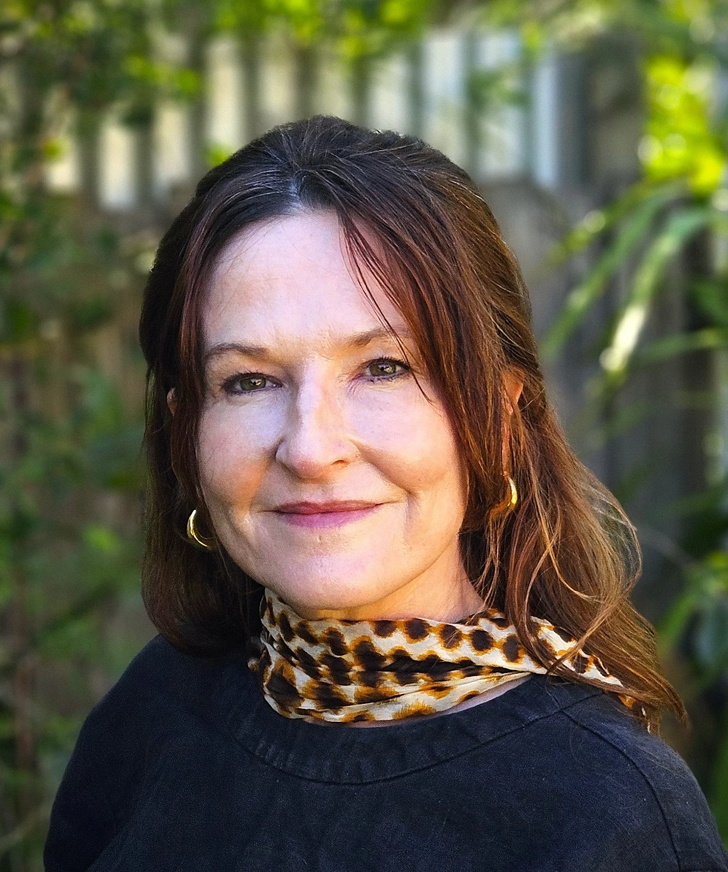 A woman with wavy brown hair and light skin smiling outdoors, wearing a black top, gold hoop earrings, and a leopard print scarf, with greenery in the background.