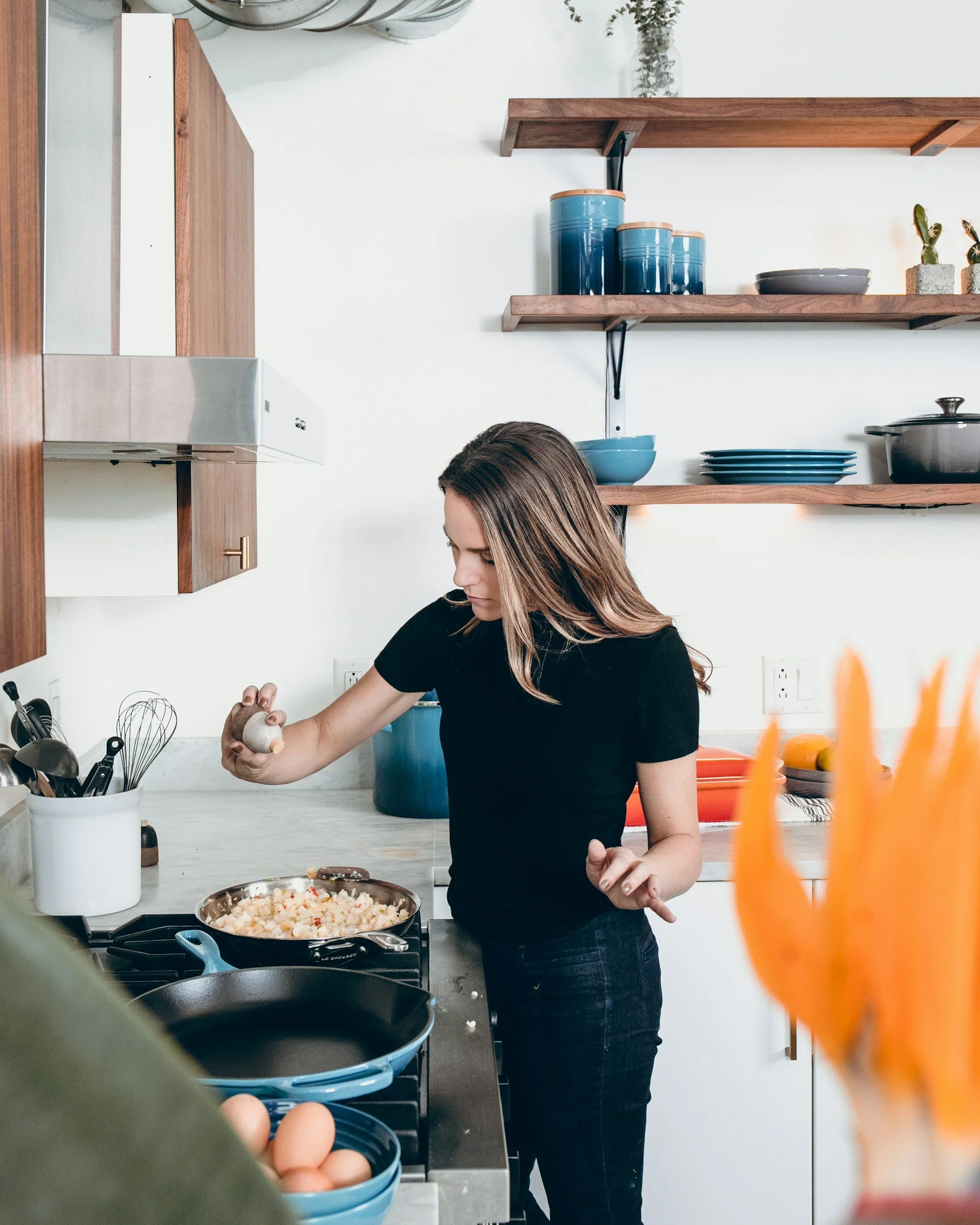 A woman cooking in a modern kitchen, stirring a dish on a stove, with shelves holding blue containers and a gray bowl on the wall behind her, and a dish of chopped vegetables in front of her.