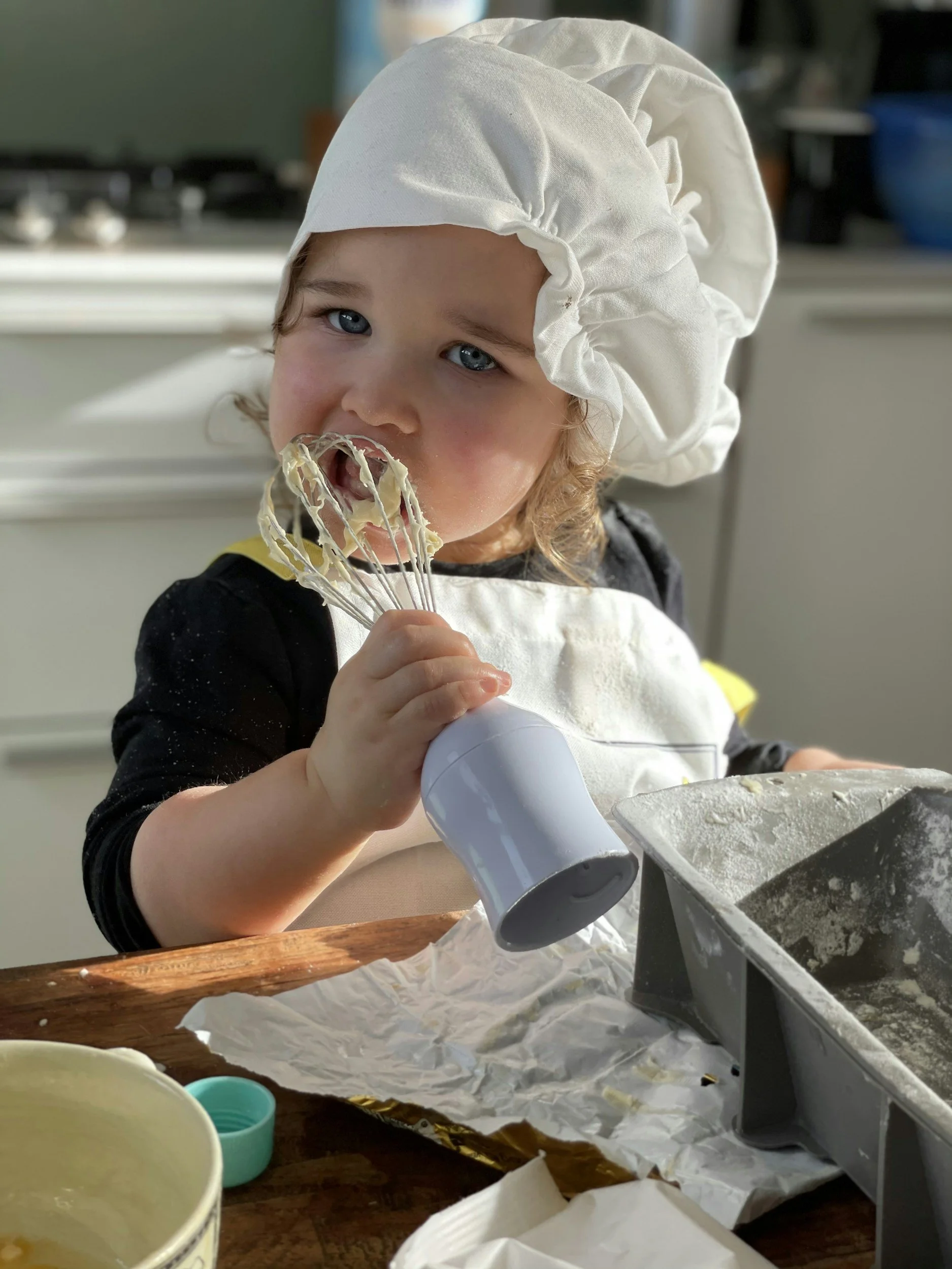 A young child with blue eyes wearing a white chef's hat and apron, using a hand mixer to beat batter in a bowl, in a kitchen setting.