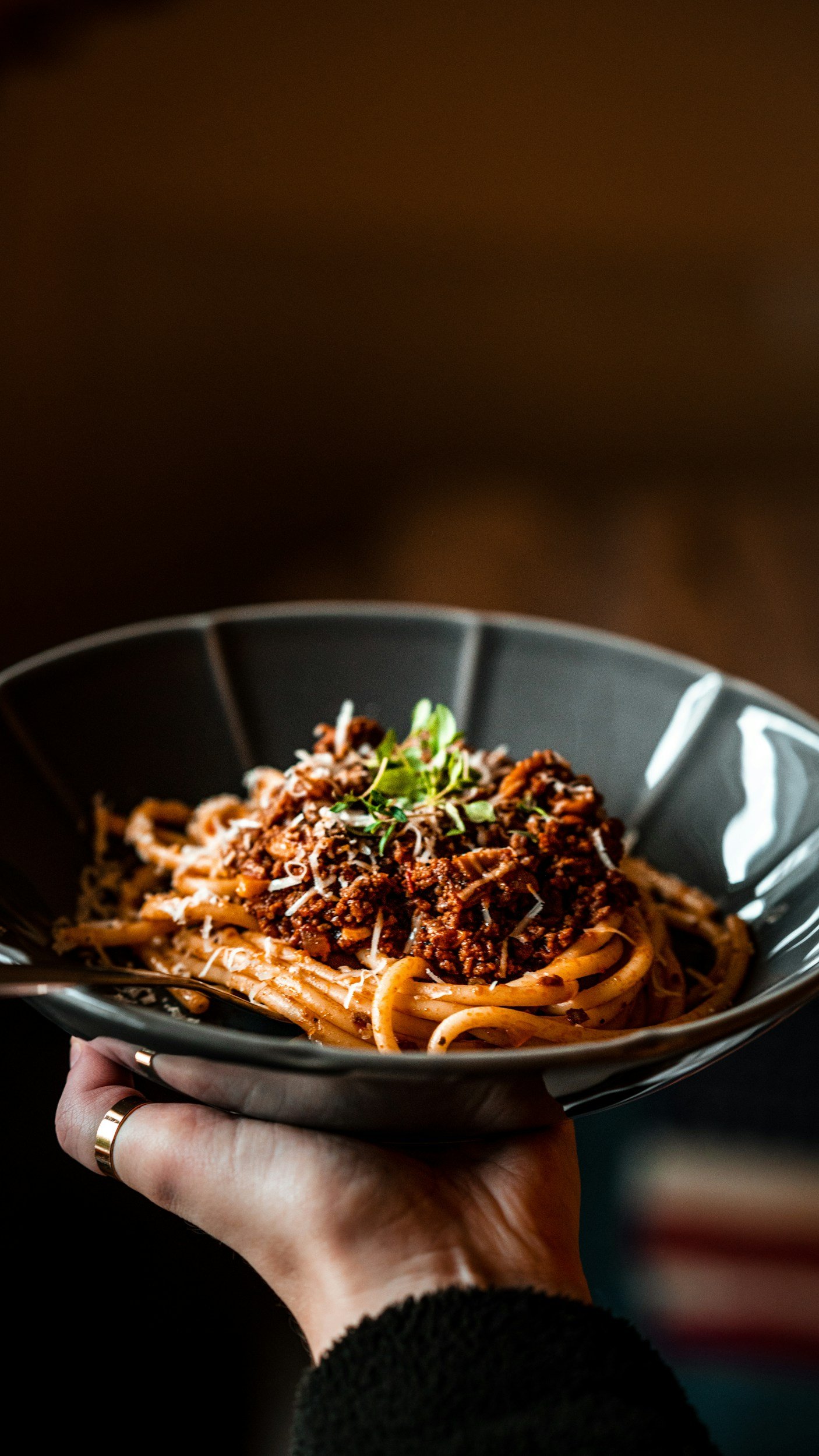 A person holding a black bowl with spaghetti topped with meat sauce, grated cheese, and a sprig of herbs.