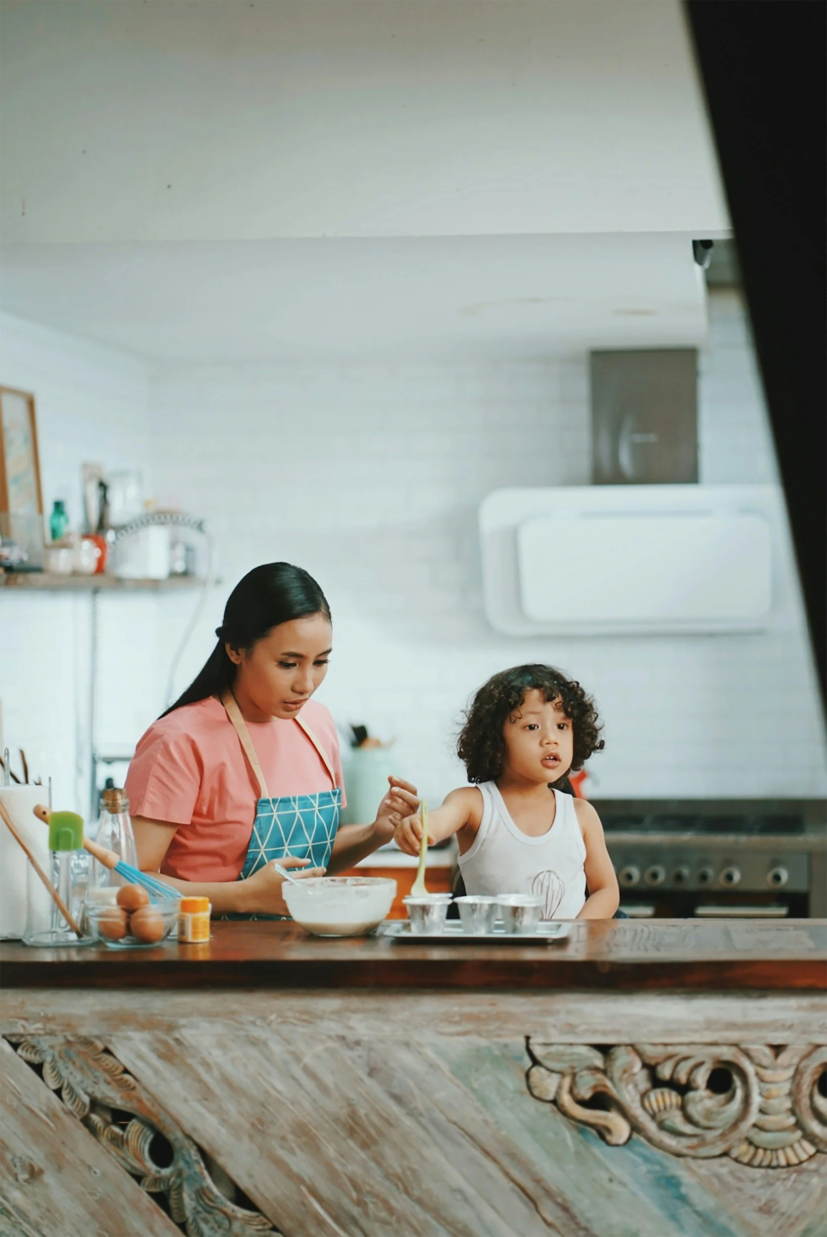 A woman and a young girl in a kitchen, preparing food together at a wooden counter with various kitchen utensils and ingredients visible.