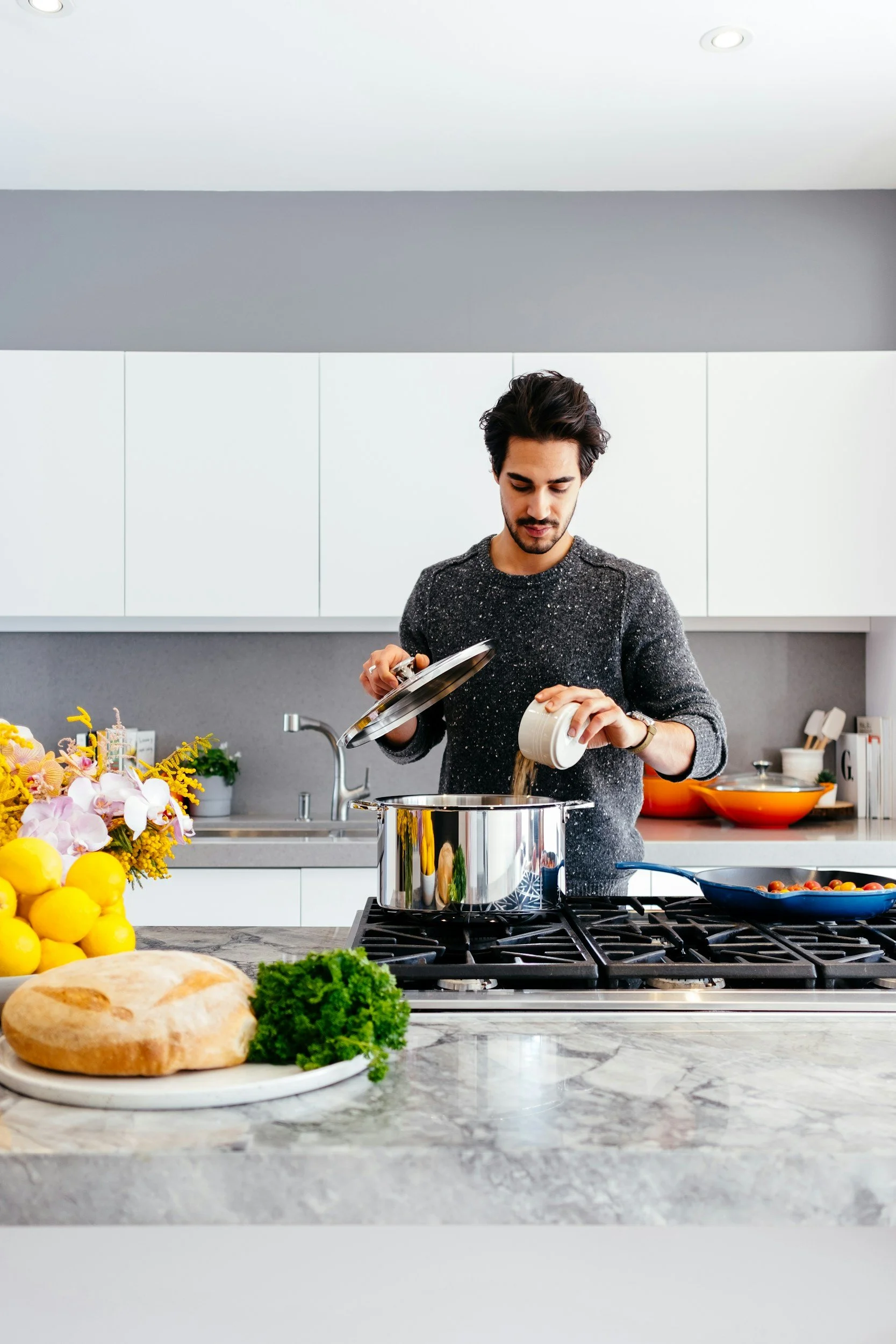 A man cooking on a stove in a modern white kitchen with fresh vegetables, a loaf of bread, flowers, and cooking ingredients on the counter.