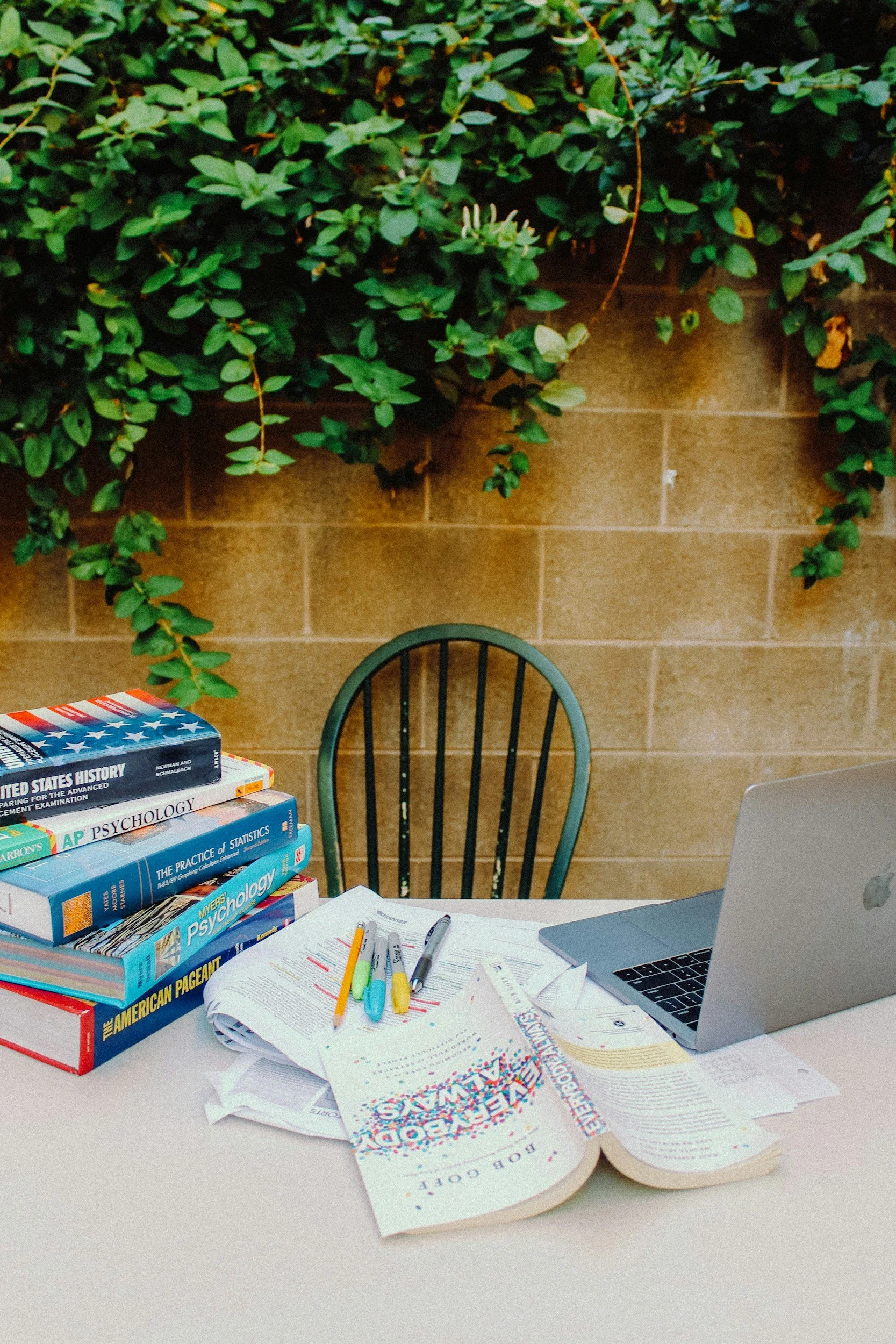 A study desk with stacked textbooks, notebooks, highlighters, pens, and a laptop, set against a wall with green leafy plants overhead.